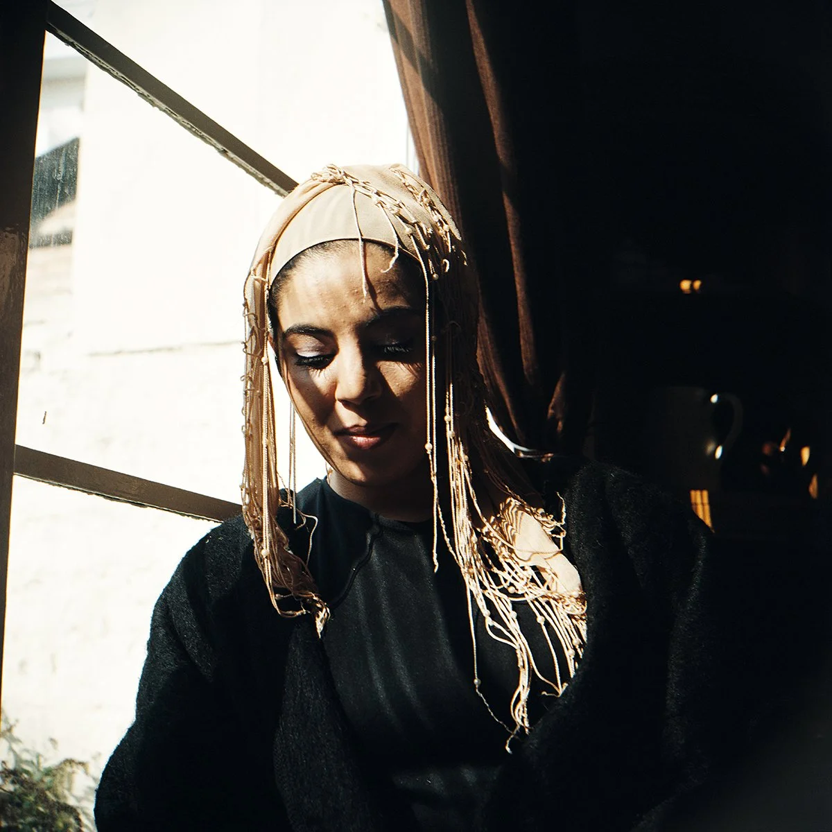 Jeune femme avec un voile beige et des perles, regardant vers le bas, dans une pièce sombre près d'une fenêtre avec rayons de soleil.