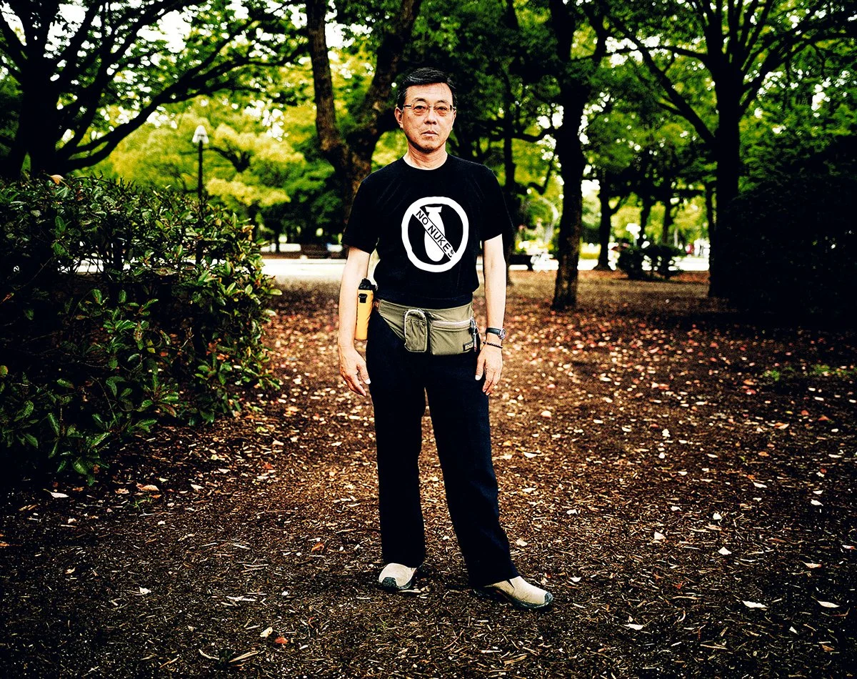 Un homme debout dans un parc avec des arbres verts en arrière-plan, portant un t-shirt noir avec un symbole anti-nucléaire et la mention "No Nukes", un sac à la taille, et une arme à côté de lui.