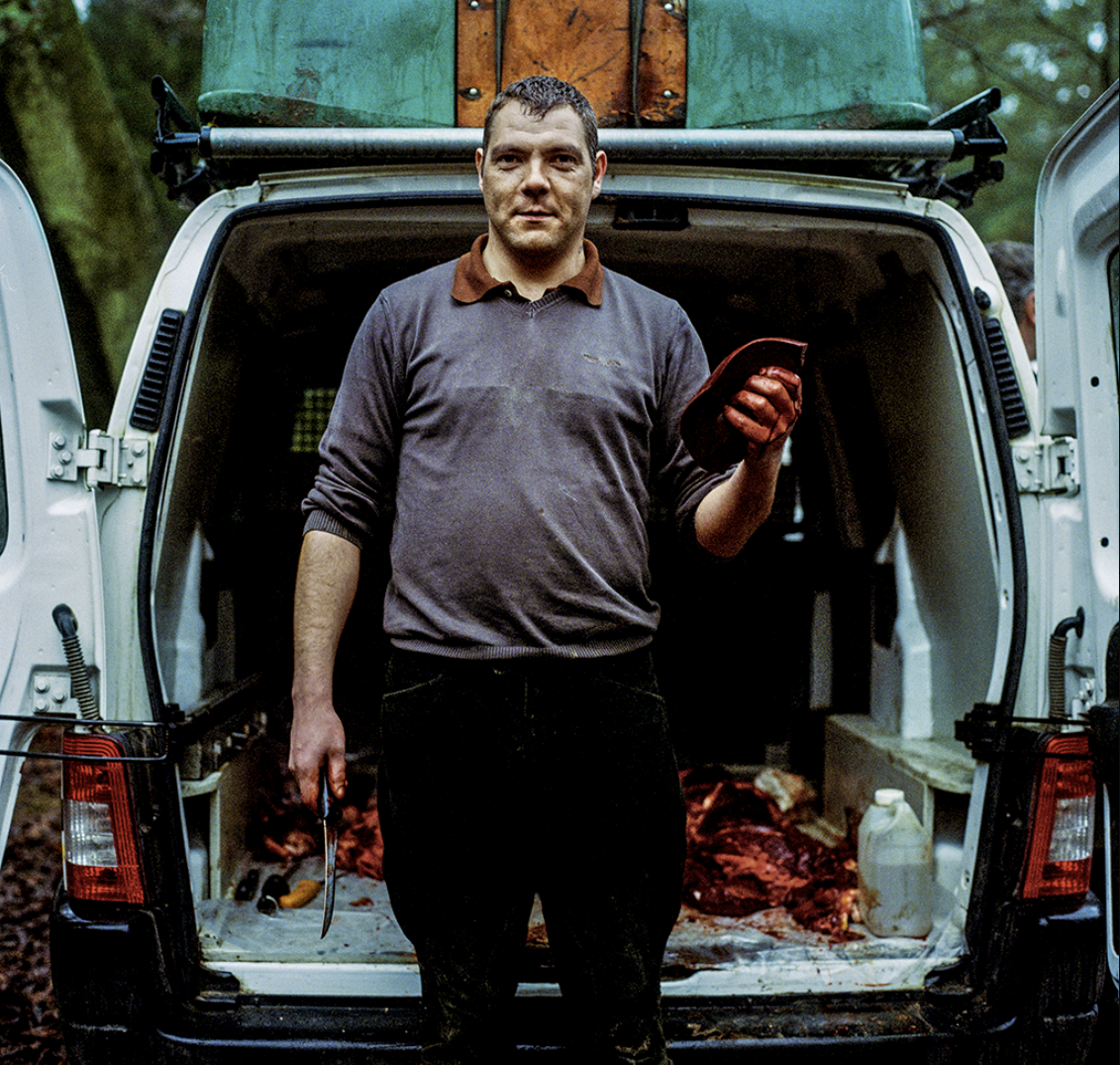 Homme debout à l'arrière d'un véhicule, tenant un couteau et un carnet rouge, portant des gants en caoutchouc rouges, dans un environnement forestier.