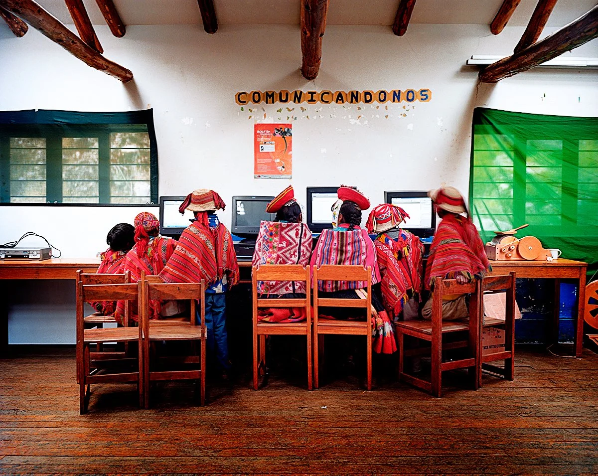 Group of people dressed in brightly colored traditional clothing, using computers in a room with a sign on the wall reading "COMUNICANDO NOS" in colorful letters.