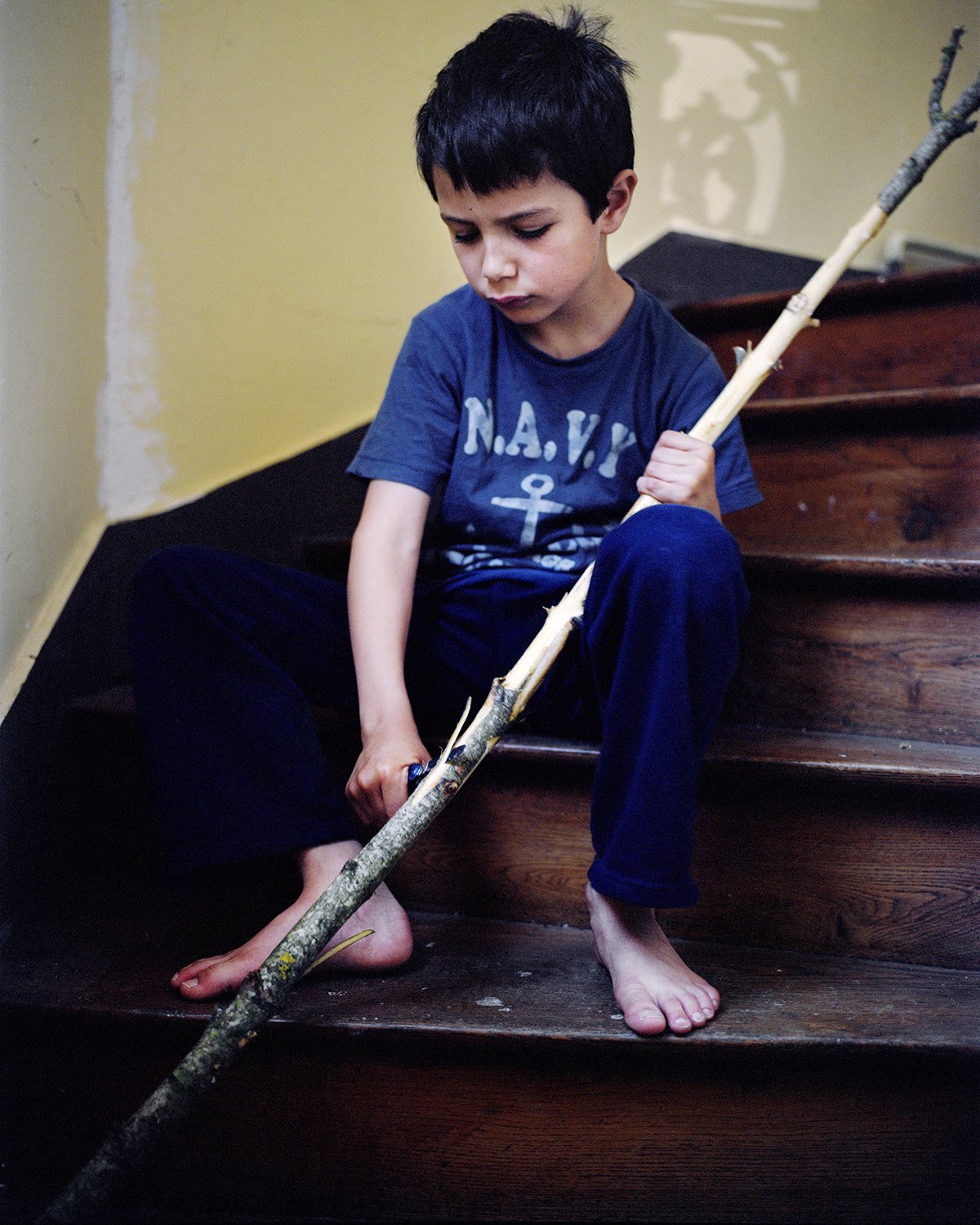 A boy sitting on wooden stairs, holding a tree branch in his hand, gazing pensively. He's wearing a blue T-shirt with an anchor and letters.