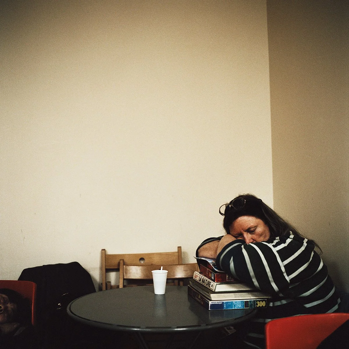 Une femme avec des lunettes sur la tête, portant un pull à rayures noires et blanches, dort avec la tête reposant sur une pile de livres sur une table ronde noire. Un petit chien est visible dans un sac à côté d'elle.