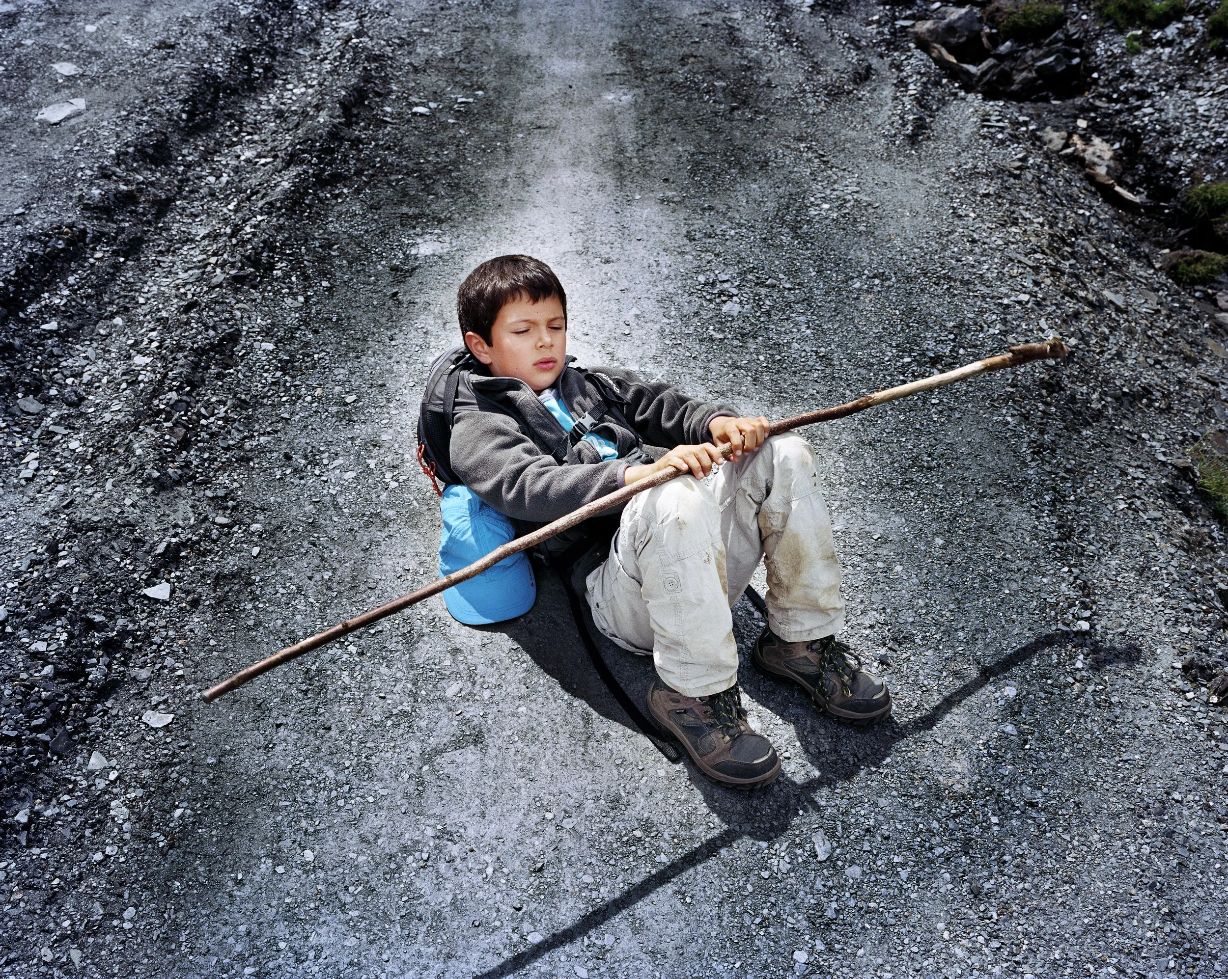 Un garçon assis sur un chemin de gravier, tenant un bâton, avec une expression concentrée.