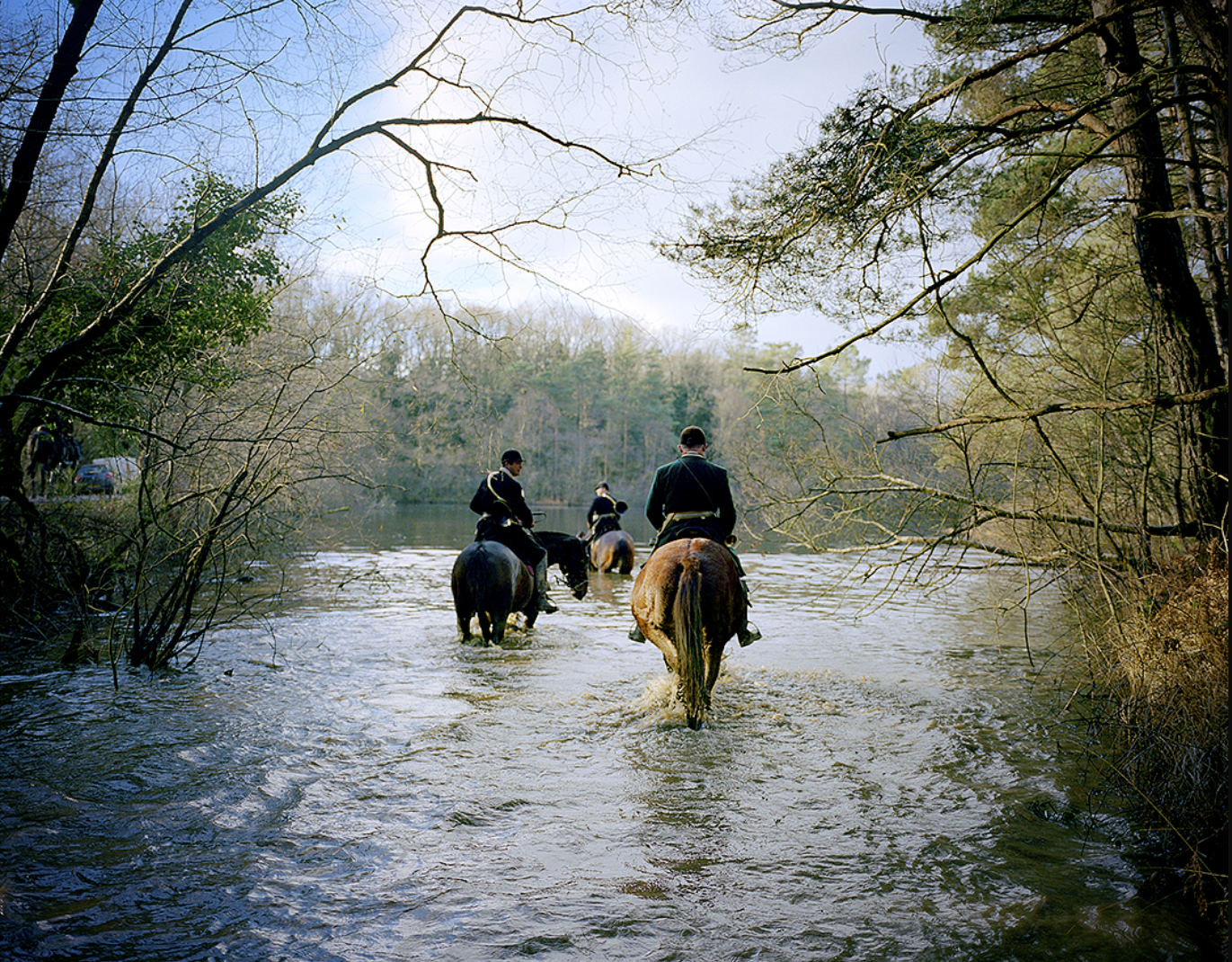 Trois cavaliers traversent une rivière en forêt, avec des arbres nus et un ciel clair en arrière-plan.