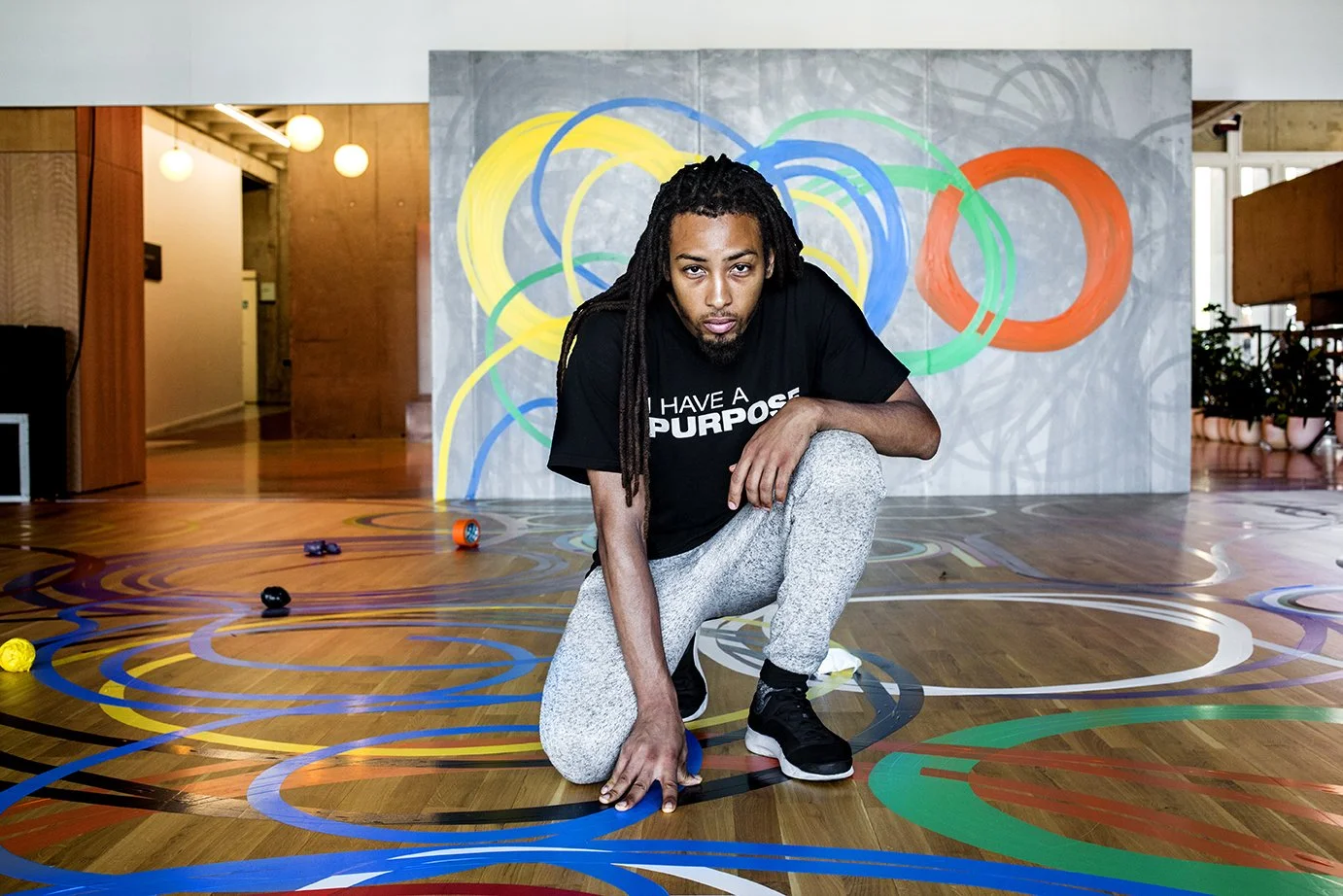 A man with dreadlocks, wearing a black T-shirt and gray jogging pants, poses in a room with a wooden floor, a wall decorated with colorful painted circles, and a few toys or objects on the floor.