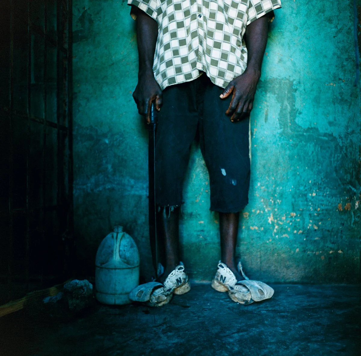 A man holds a drink can in one hand. He's wearing worn-out clothes, torn black shorts, worn sneakers, and is standing against a turquoise wall. There's a bottle on the floor beside his feet.