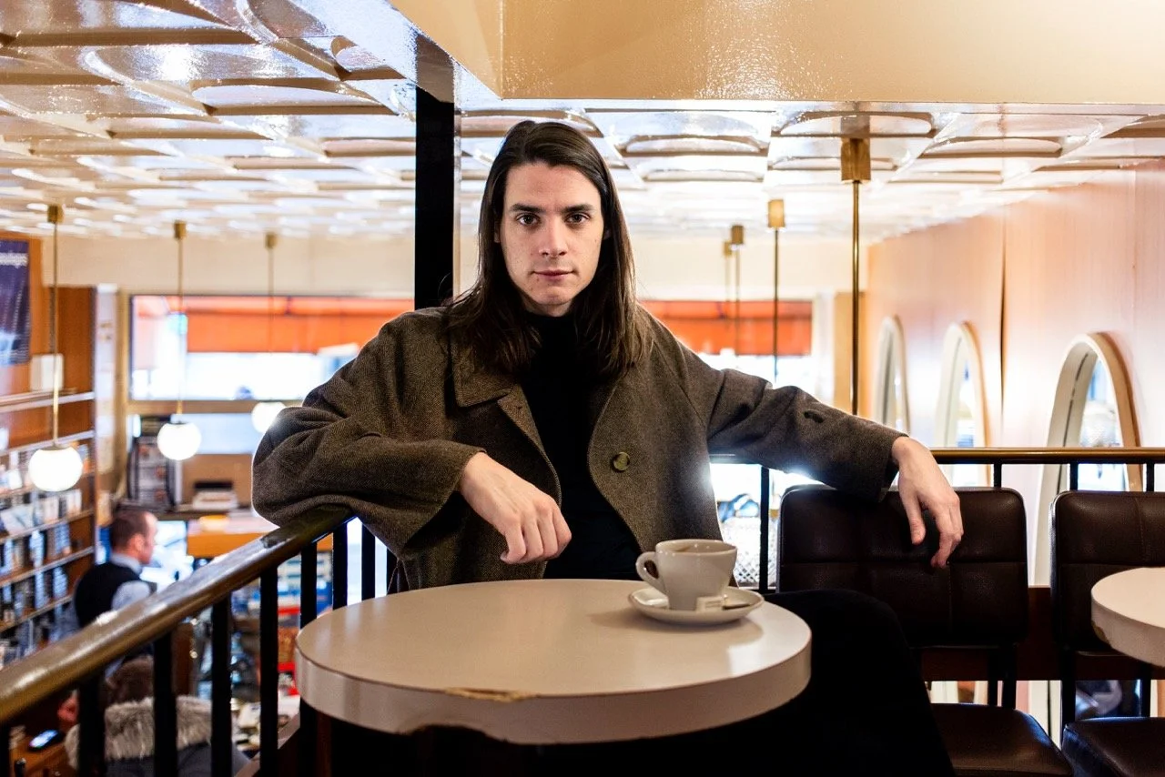 Young man with long hair, seated at a table in a café, with a cup and saucer in front of him, in a retro setting with a cube-panelled ceiling.