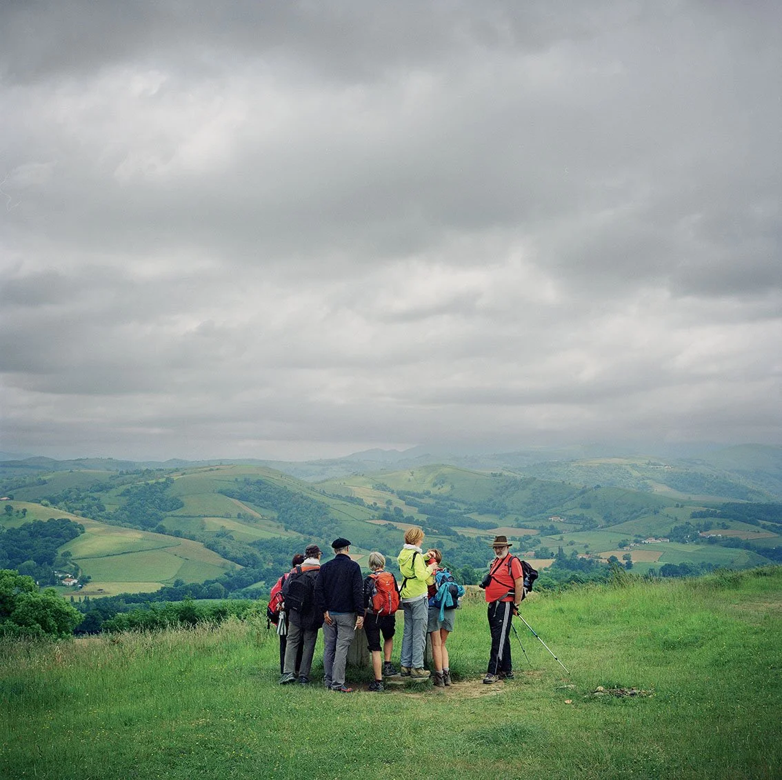A group of hikers in the open air on a hill with a green landscape and distant mountains under a cloudy sky.