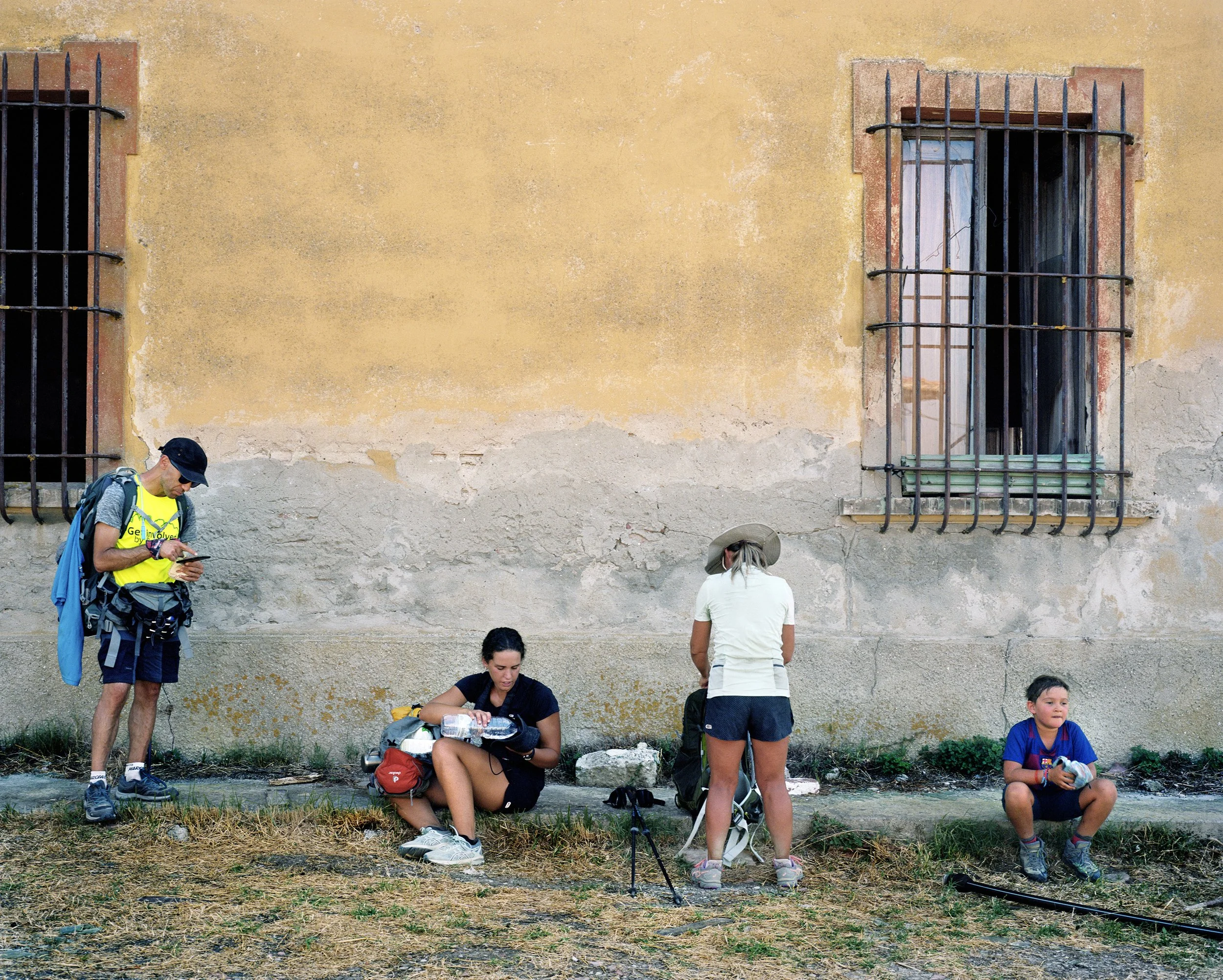 Group of people sitting and standing in front of an ancient wall with barred windows, some listening or using a telephone, casually dressed with bags and hiking gear.