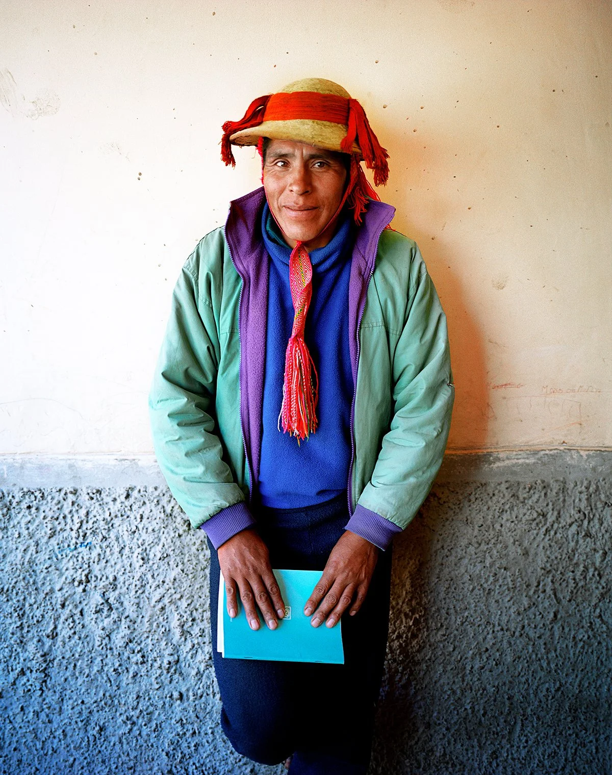 A man wearing a colorful hat, blue sweatshirt, green jacket and holding blue documents, standing against a white wall with a concrete lower section.