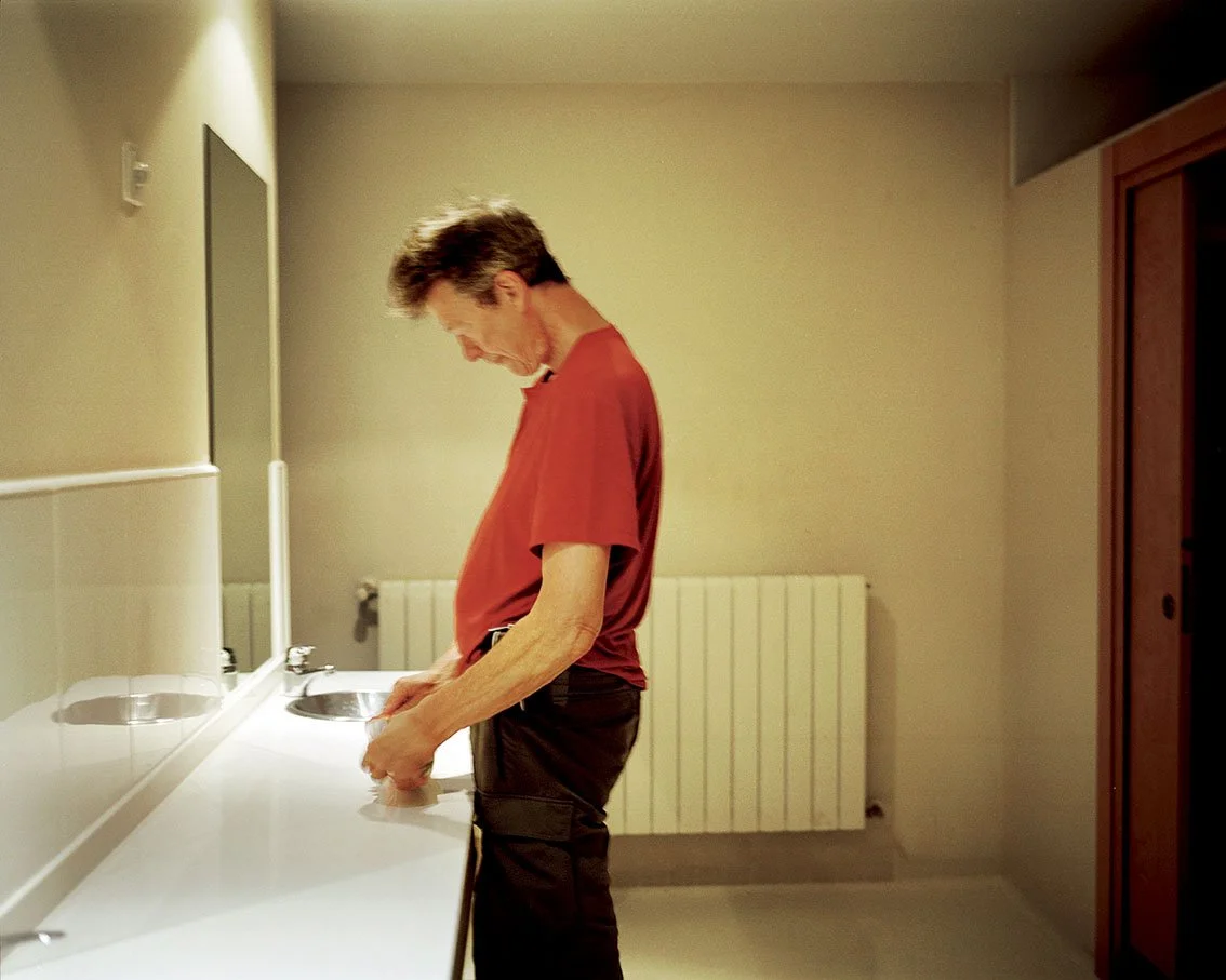 A man in a red T-shirt washes his hands at a sink in a bathroom or changing room.