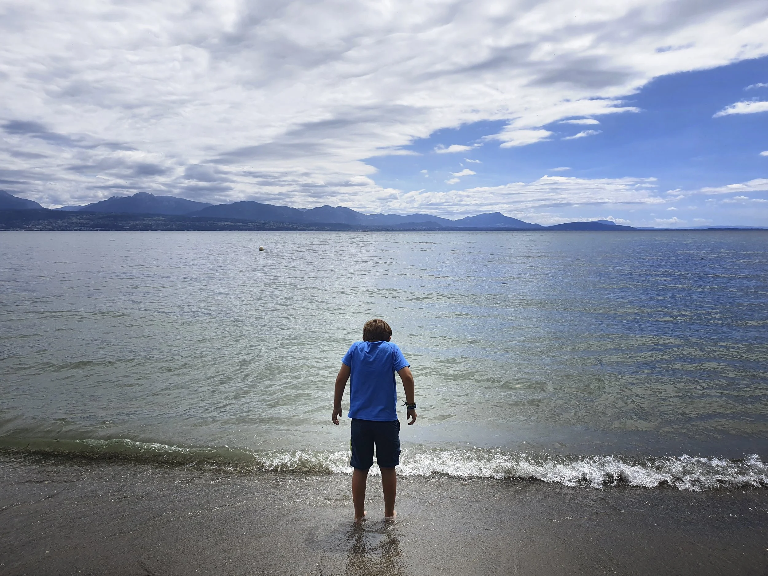 A boy standing on the beach looks out over the expanse of a lake with mountains in the background, the sky partly cloudy.