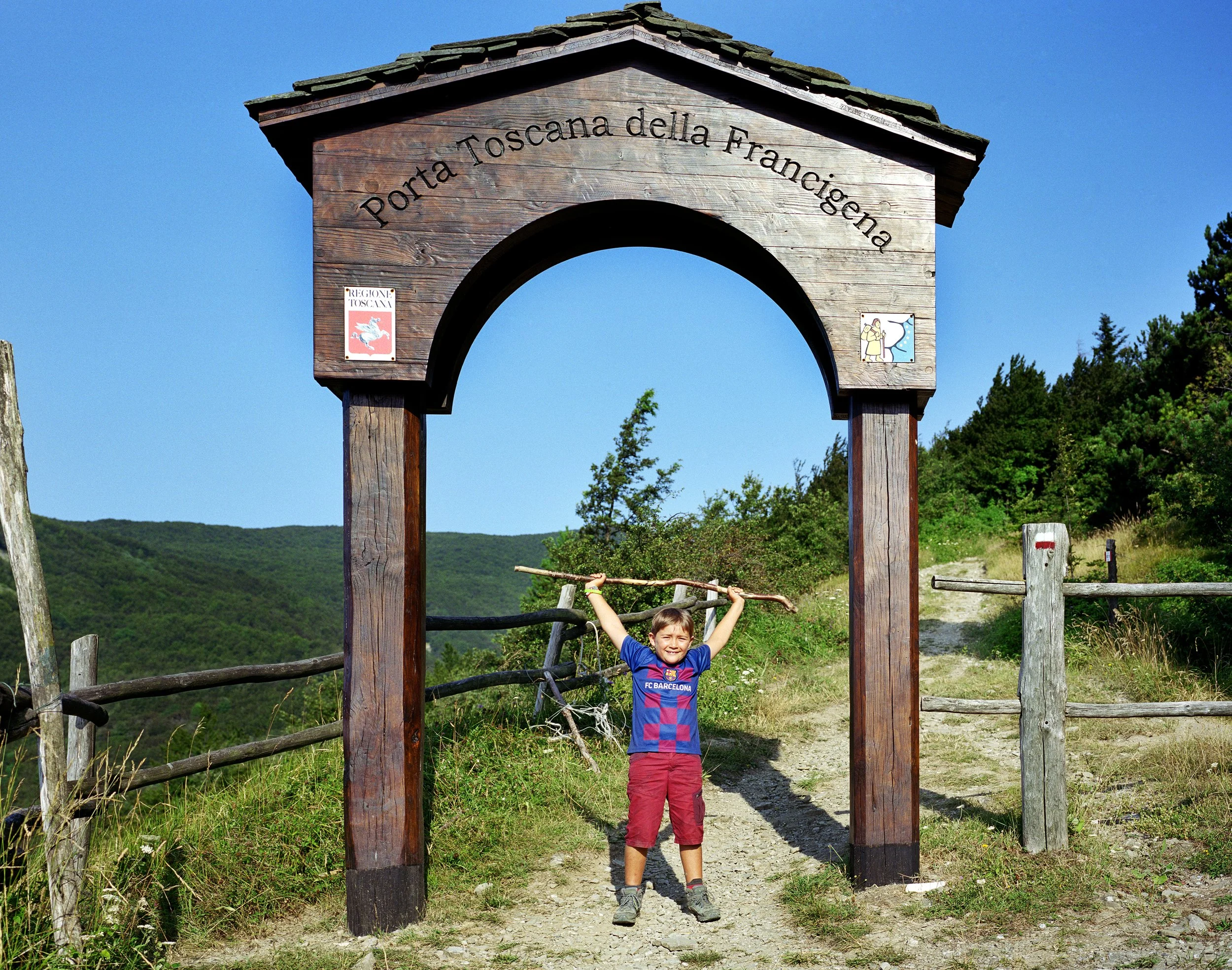 A smiling boy in an FC Barcelona T-shirt, carrying a branch as a staff above his head, stands under a wooden arch marked "Porta Toscana della Fraternità" in a verdant mountain region of Italy.
