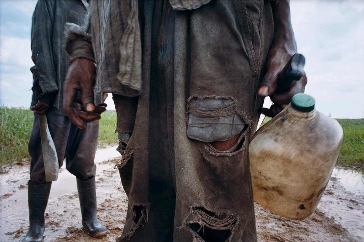 Two people in a field, wearing worn clothing and work clothes, with rubber boots, one holding a beige plastic bottle.