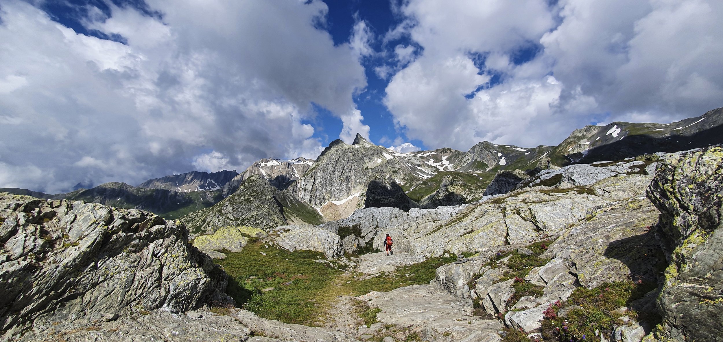 Hiker with a red backpack walking on a rocky path in a mountain landscape with snow-capped peaks, partly cloudy sky