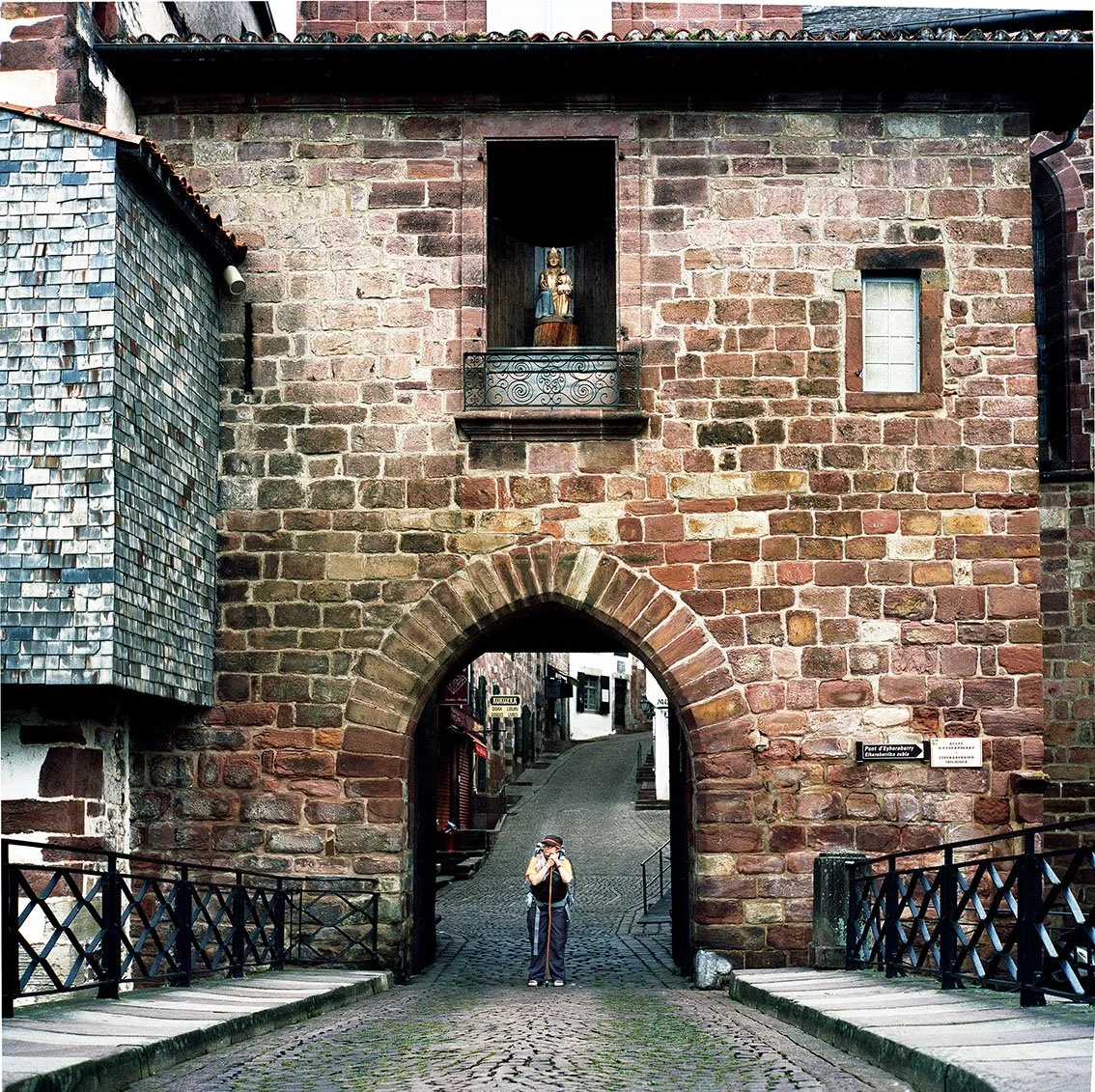 An arched doorway in a red stone wall, with a person taking a photo in front of it, and buildings in the background.
