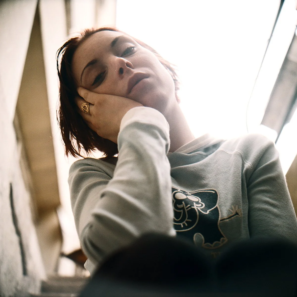 Jeune femme avec brune aux cheveux courts, portant un sweat gris, regardant vers le haut, avec la main sur la tête, à l'intérieur près d'une fenêtre lumineuse