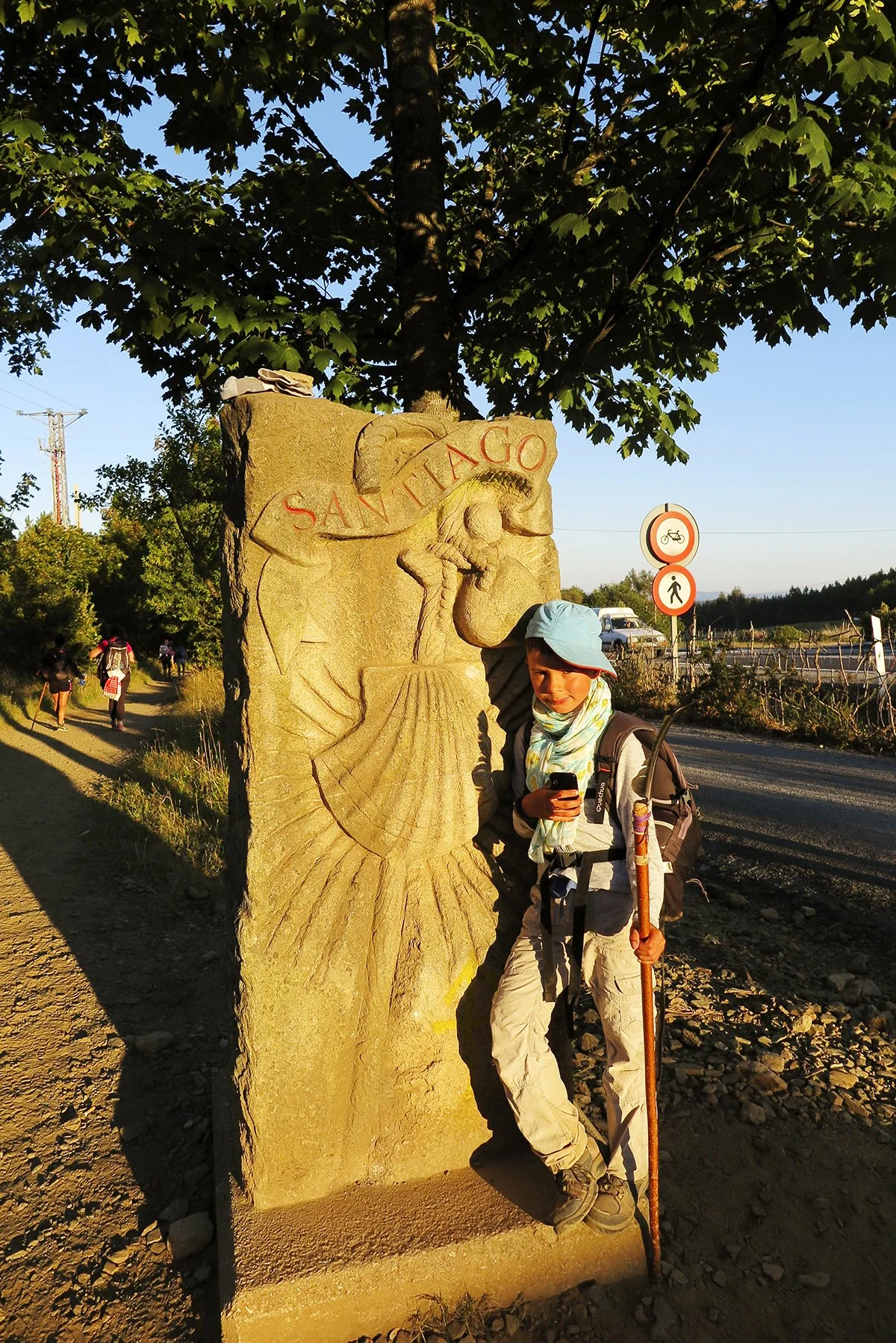 A woman standing next to a stone sculpture depicting a human face and an animal face, with the inscription "SANTIAGO" at the top, under a tree in the open air, at sunset.