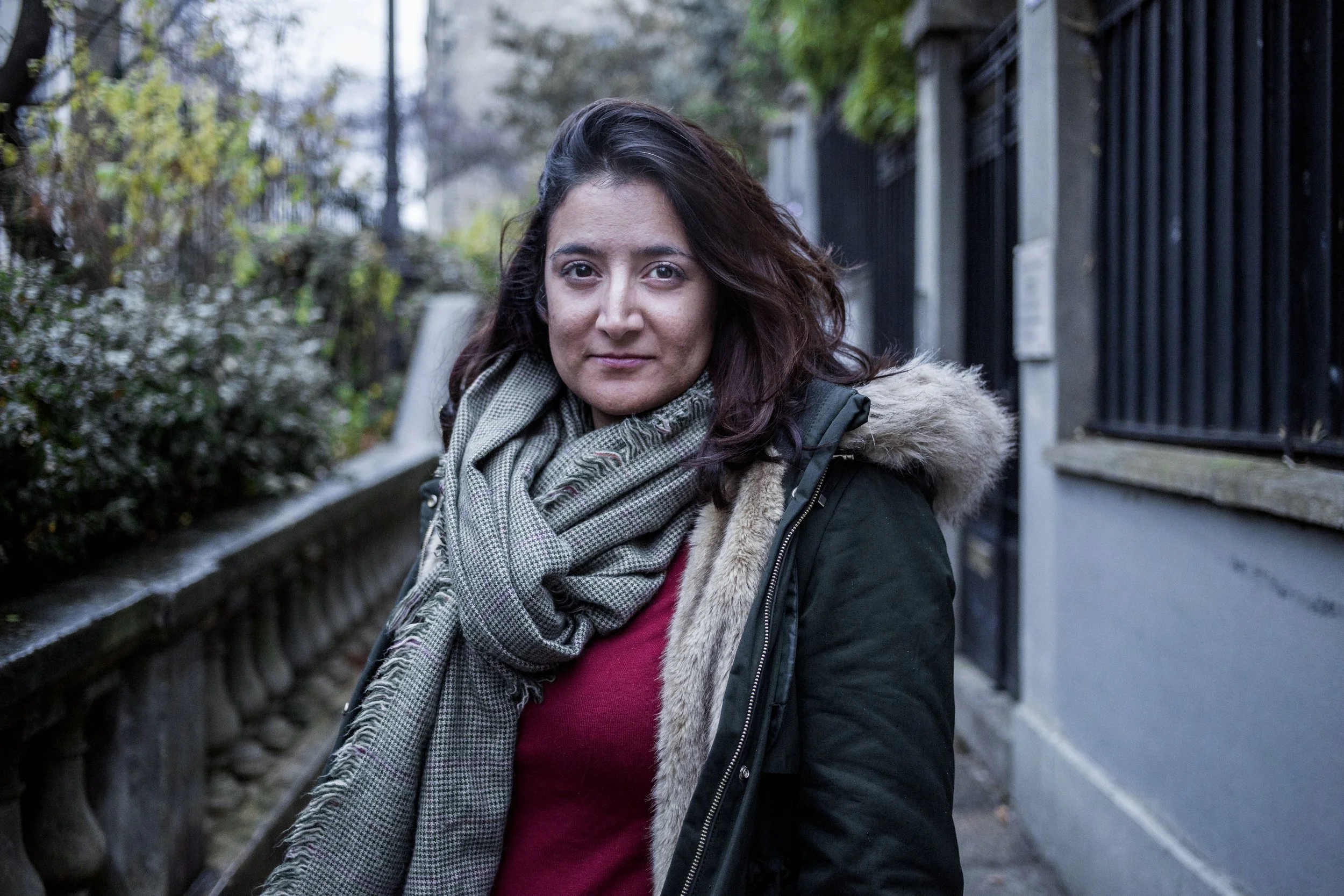 Woman with hooded coat tucked in, gray scarf, brown hair, looking at camera, outdoors in an urban environment
