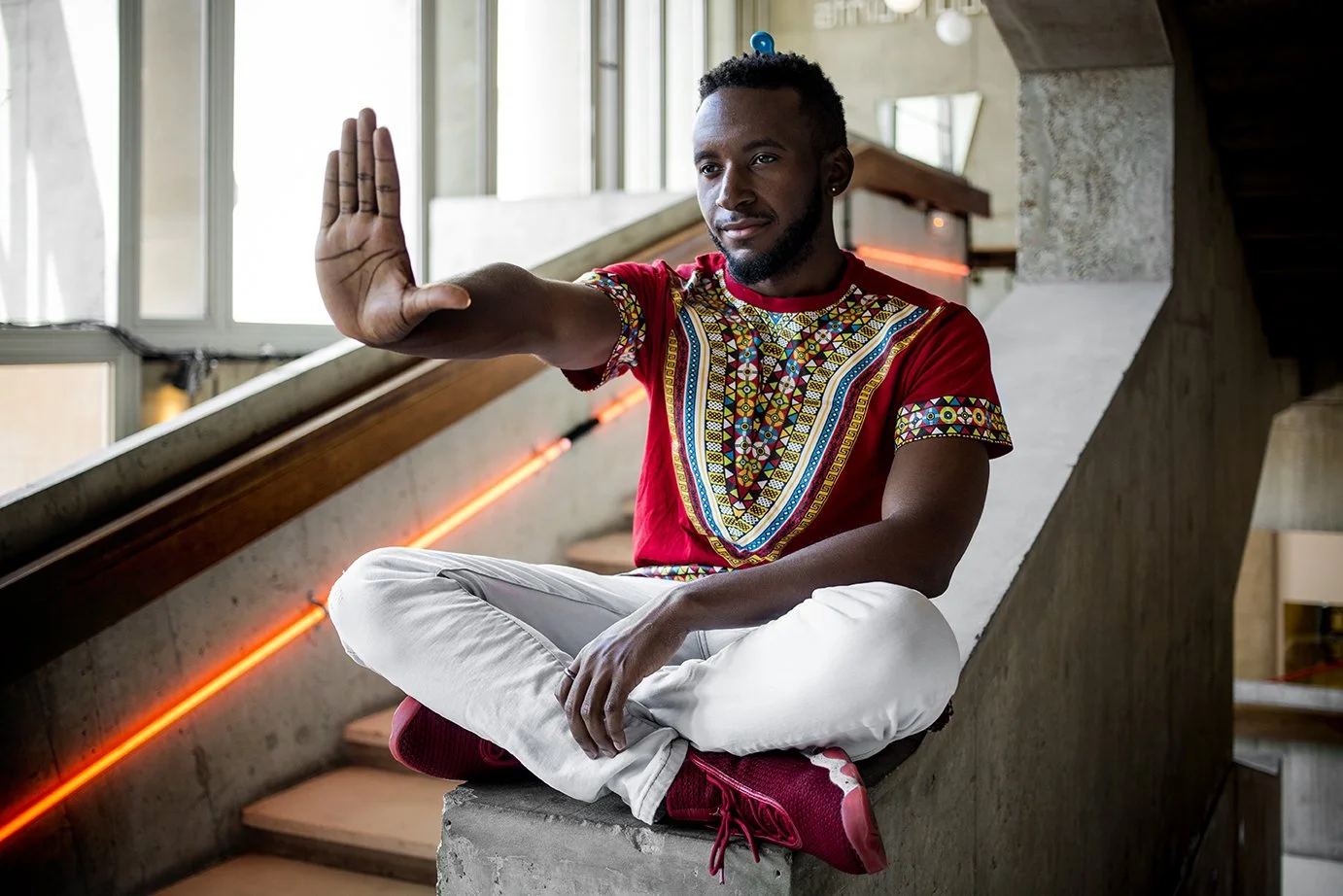A man sitting in a meditative position, wearing a colorful African garment, in a modern interior with concrete stairs and neon lighting"}.