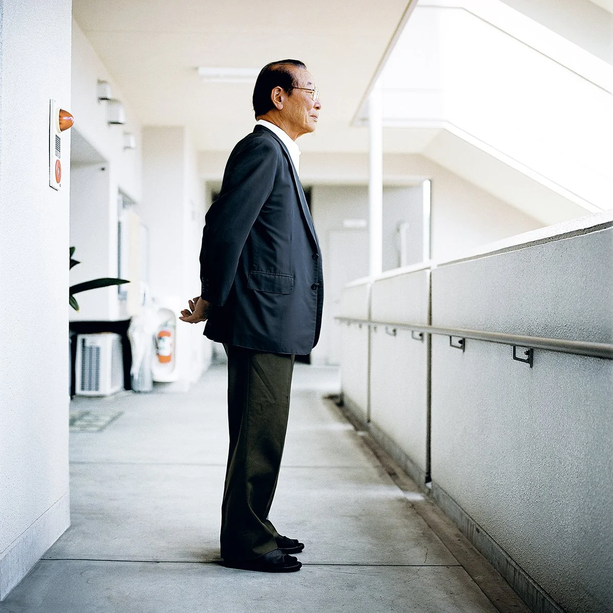 Un homme d'âge avancé portant un costume décontracté, debout dans un couloir lumineux et moderne, regardant vers la droite avec une posture relaxée.
