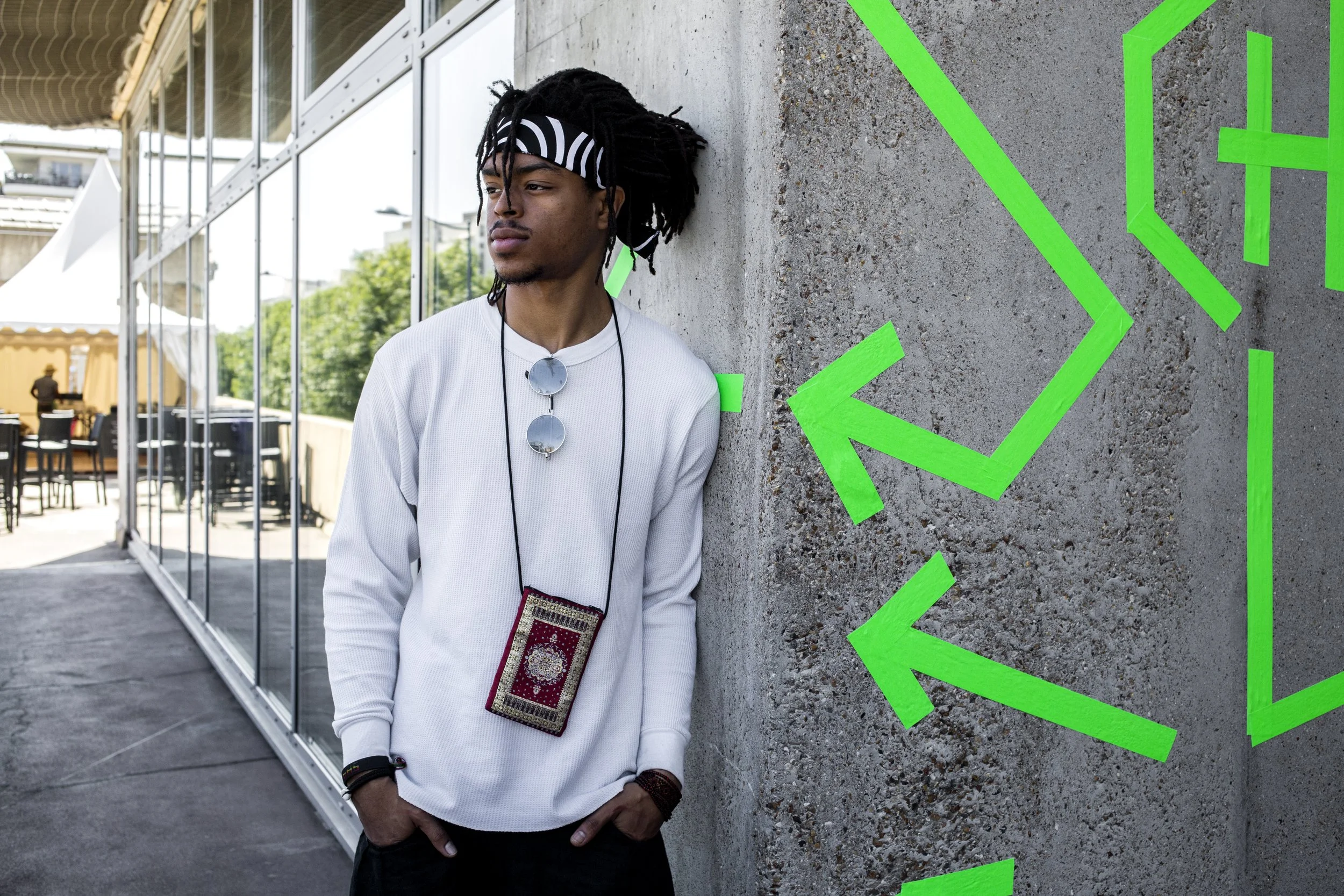 Young man wearing a white jersey, black and white headband, sunglasses hanging around his neck, and a wallet hanging around his neck, leaning against a concrete wall with green arrows in the background, outdoors.