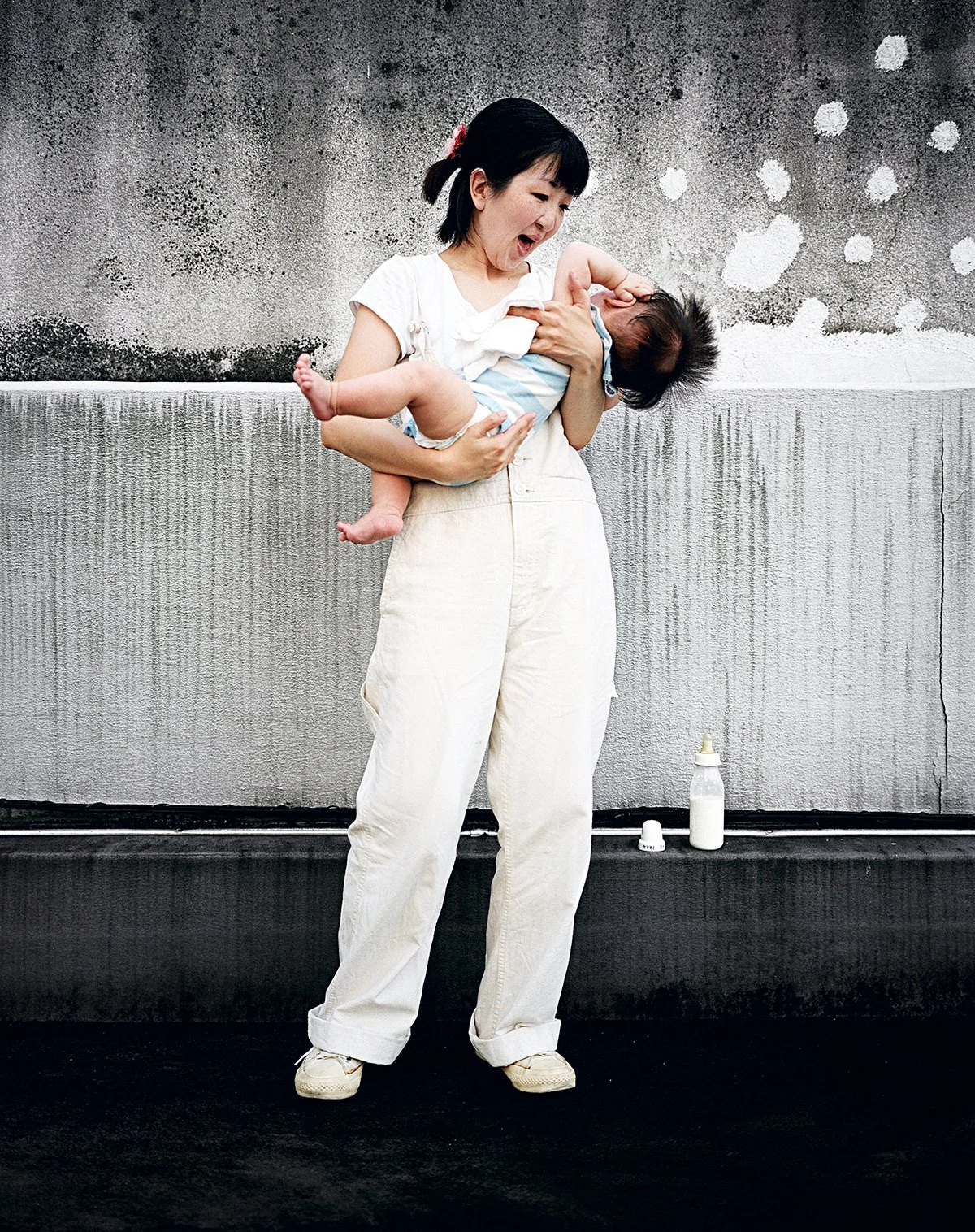 Une femme asiatique tenant un bébé en souriant et riant contre un mur gris avec des taches blanches.