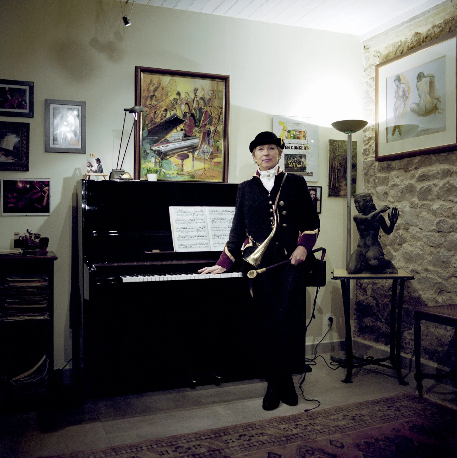 A woman dressed in a black suit with a small black hat, holding a live flute next to a black piano in a room decorated with artwork and sculpture.