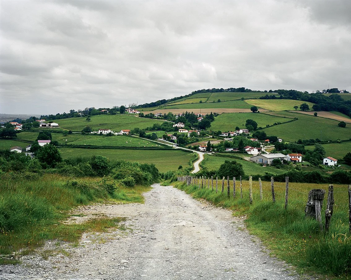 Dirt road leading to a landscape of green hills with scattered houses, under a cloudy sky.