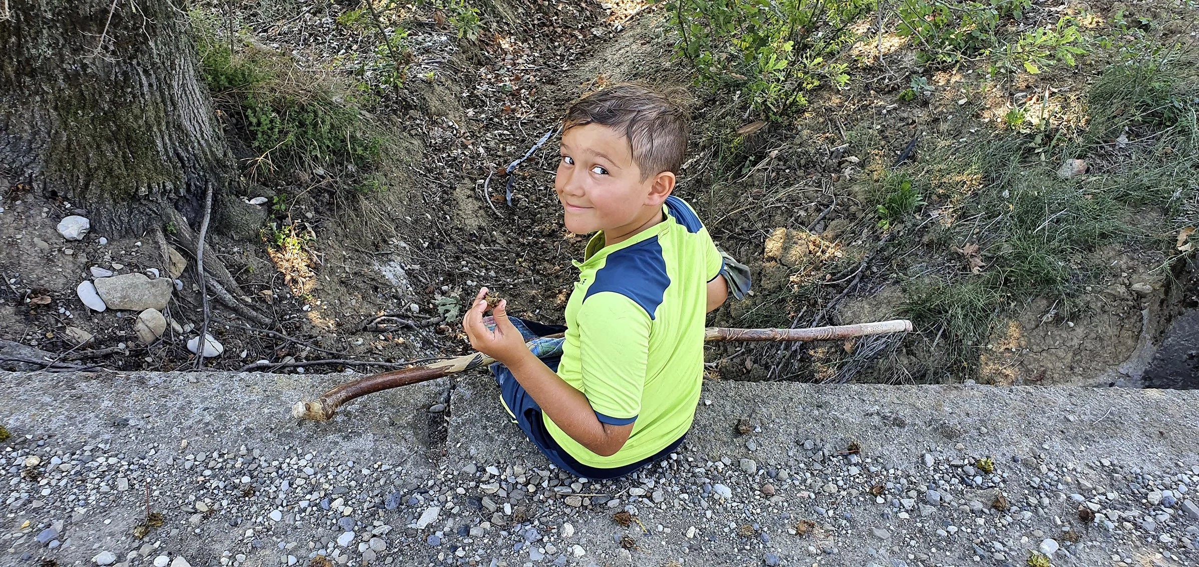 Young boy sitting on a stone platform at the foot of a tree in nature, holding a stick, looking up and smiling.
