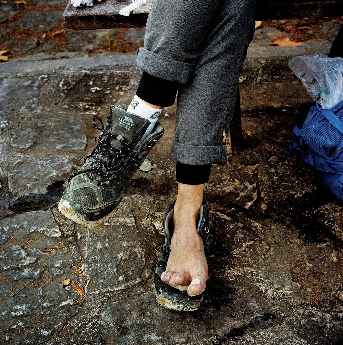 Person in hiking clothes, wearing a hiking boot on one side and a sandal on the other, sitting on rocky ground. New hiking boot on left, bare left foot with sandal on right. Blue backpack beside.