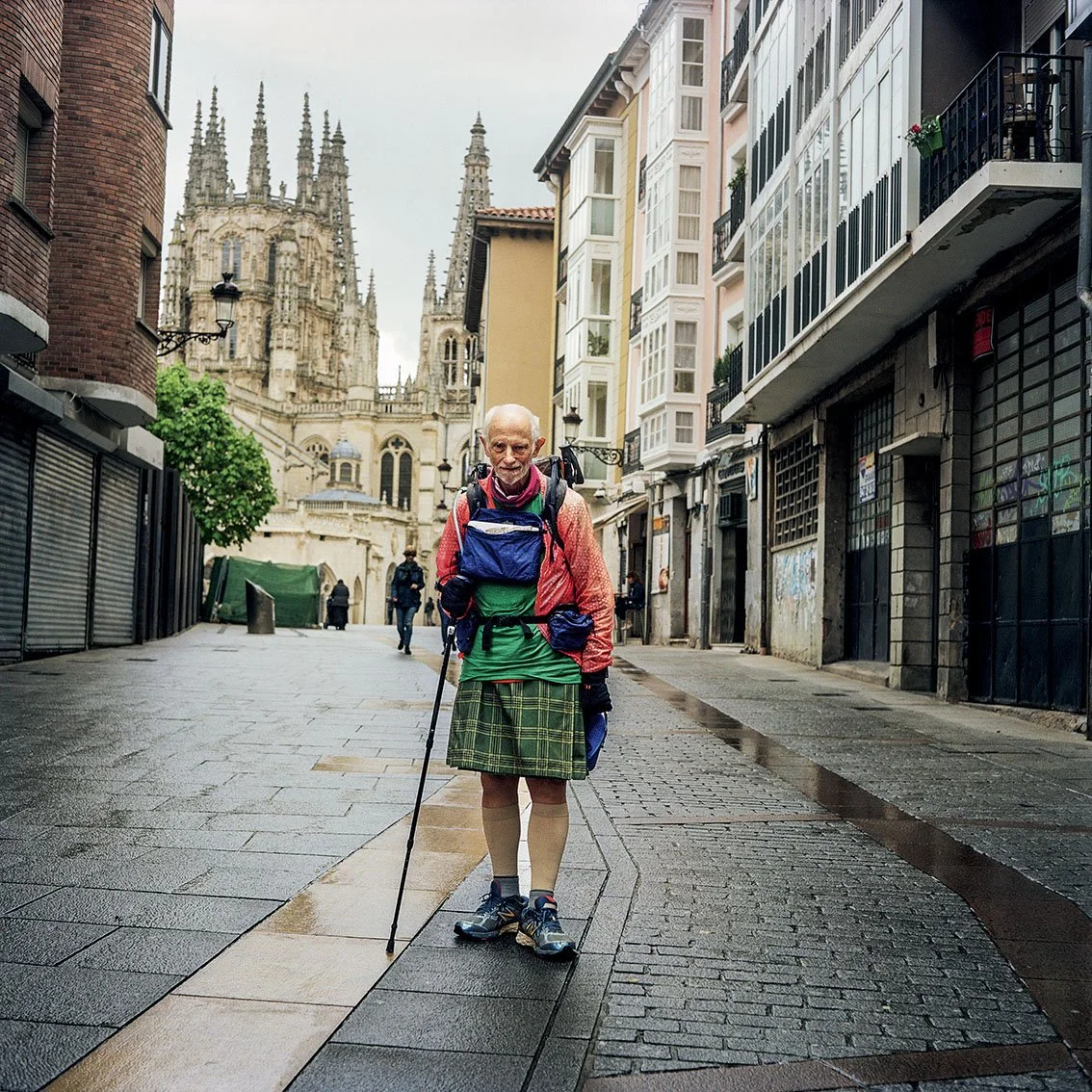 An elderly man in a tartan kilt, wearing a rucksack, pink jacket and hiking boots, walks with a cane down an empty cobbled street, with a large cathedral in the background.