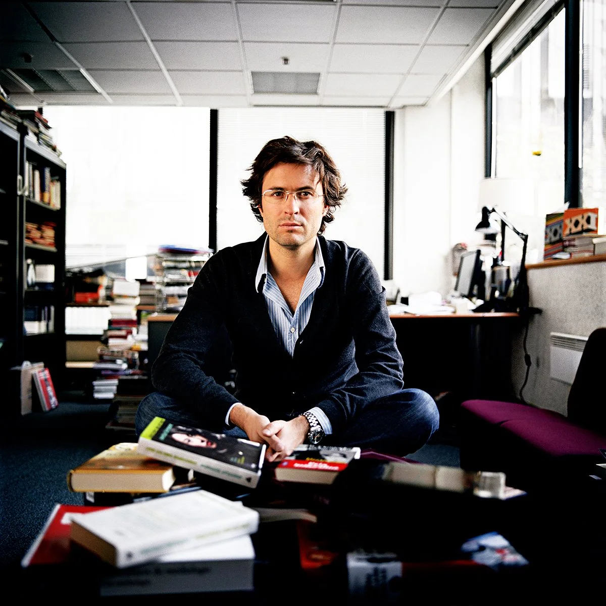 A man sitting on the floor in a cluttered office with lots of books and papers, wearing a black sweater and glasses, looking into the lens.