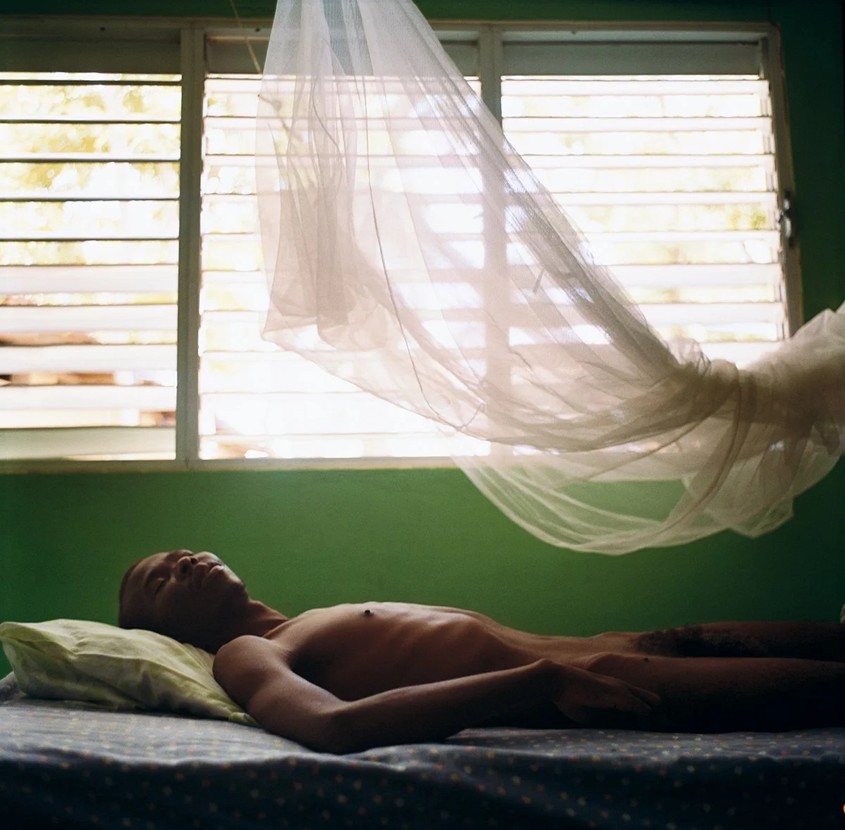 A man lies on a bed, unclothed, in a room with green walls and a window with wooden shutters, a white drapery floating above him.