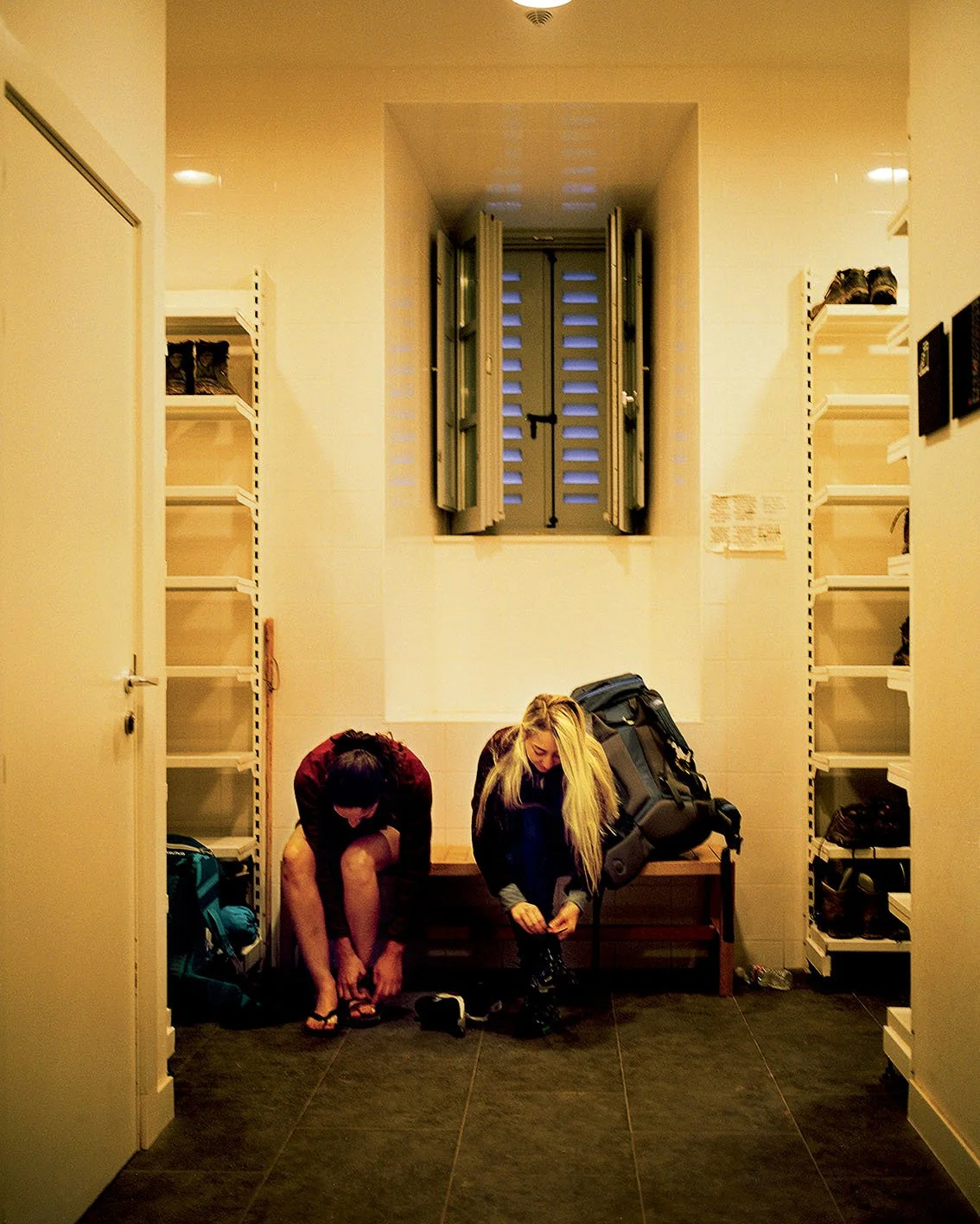 Two women in the doorway of a refuge, one getting ready to put on her shoes and the other tying her shoelaces, with rucksacks beside them.