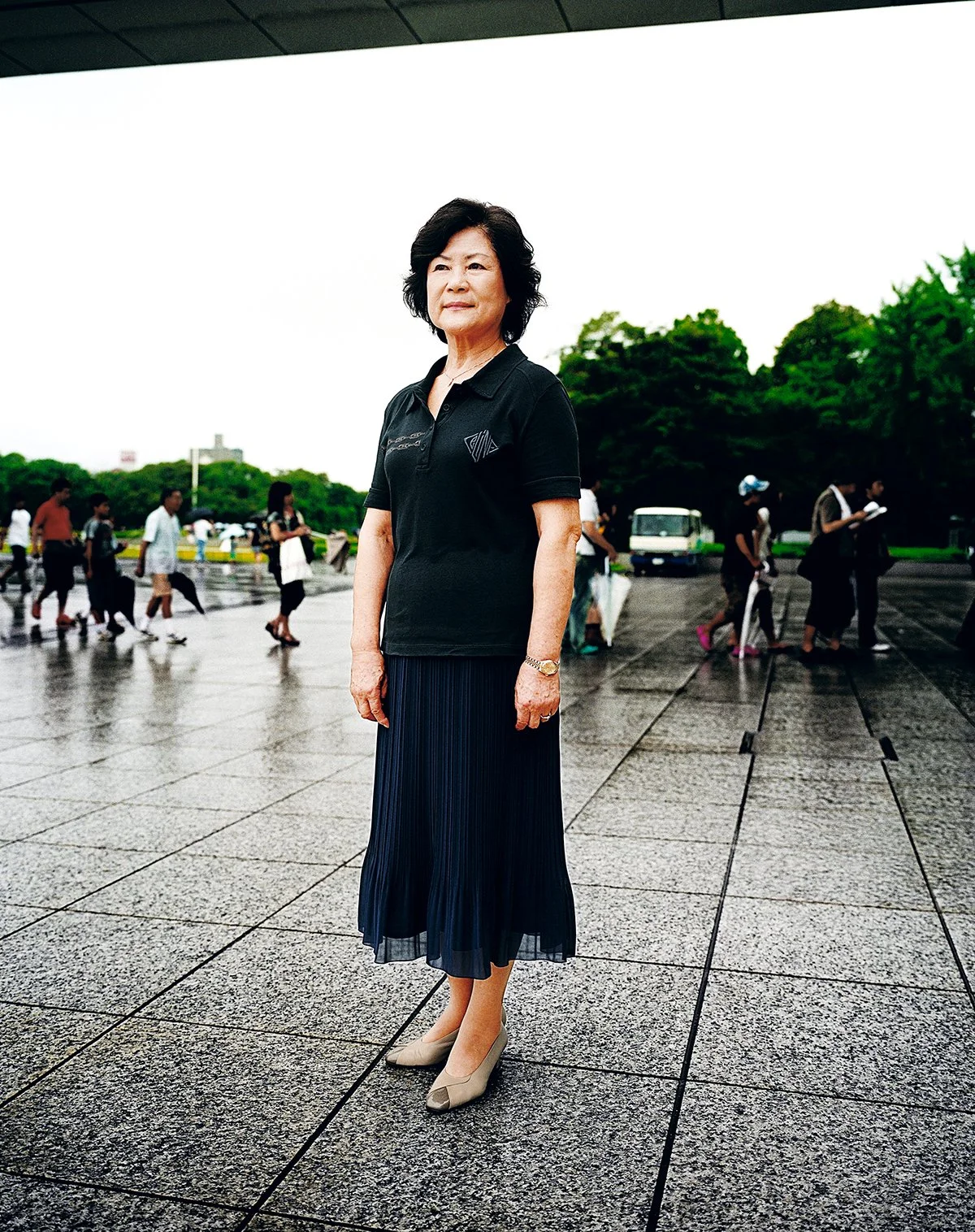 Une femme âgée debout sur un sol pavé, portant une robe longue bleu et un blazer noir, avec un groupe de personnes et des arbres en arrière-plan.