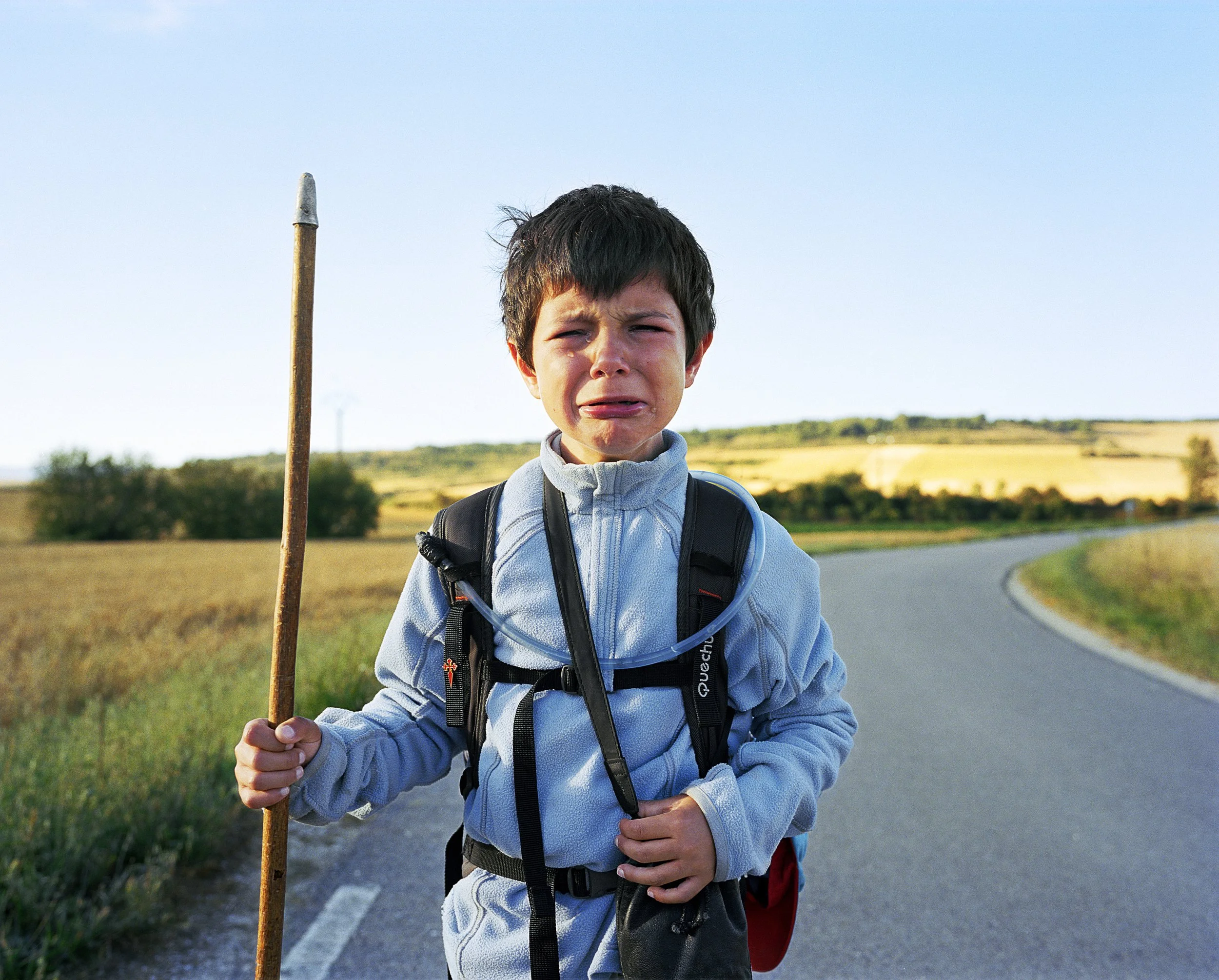 Un jeune garçon pleure en tenue de marcheur avec un sac à dos sur une route de campagne ensoleillée.