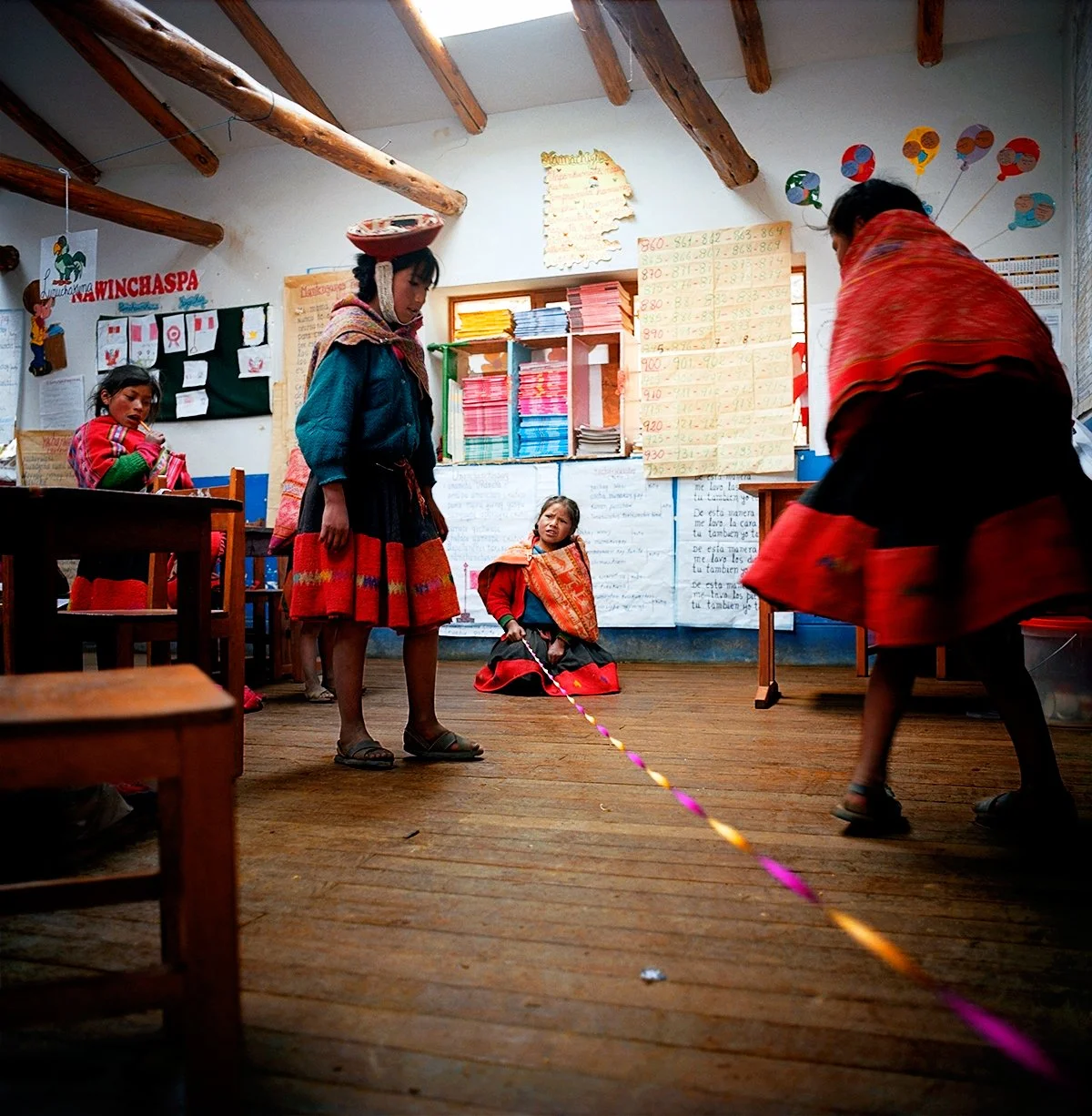 Andean children play skipping rope in a school classroom decorated with posters and bright colors.