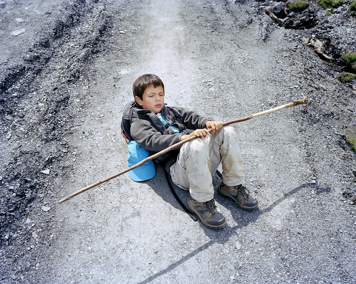 A boy sitting on the ground on a gravel path, holding a long branch like a walking stick, with a backpack, in a natural setting.