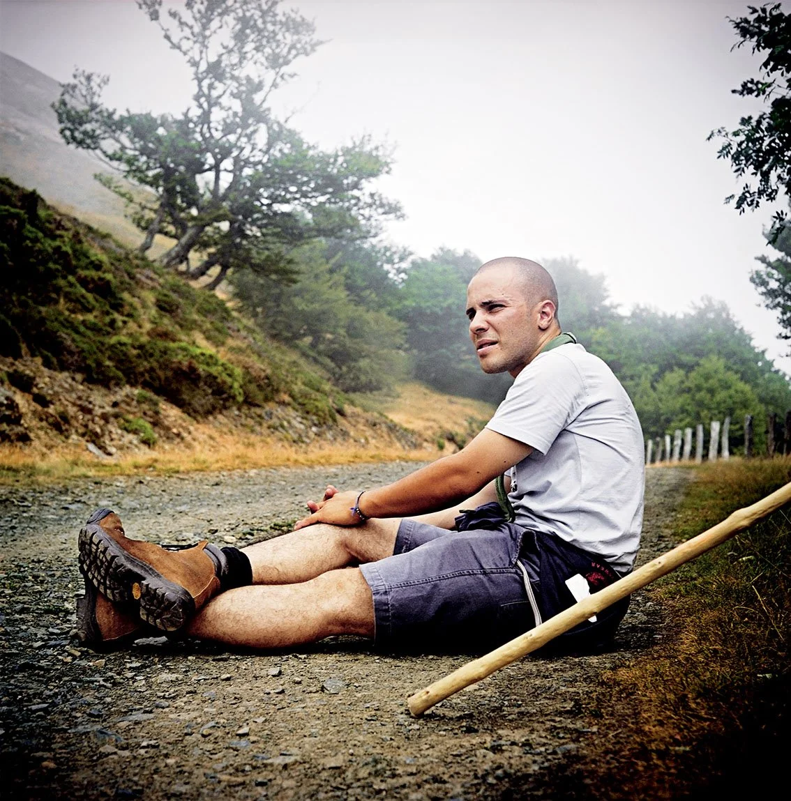Man sitting on the ground on a hiking trail in wooded surroundings, looking tired or injured, with a walking stick beside him.