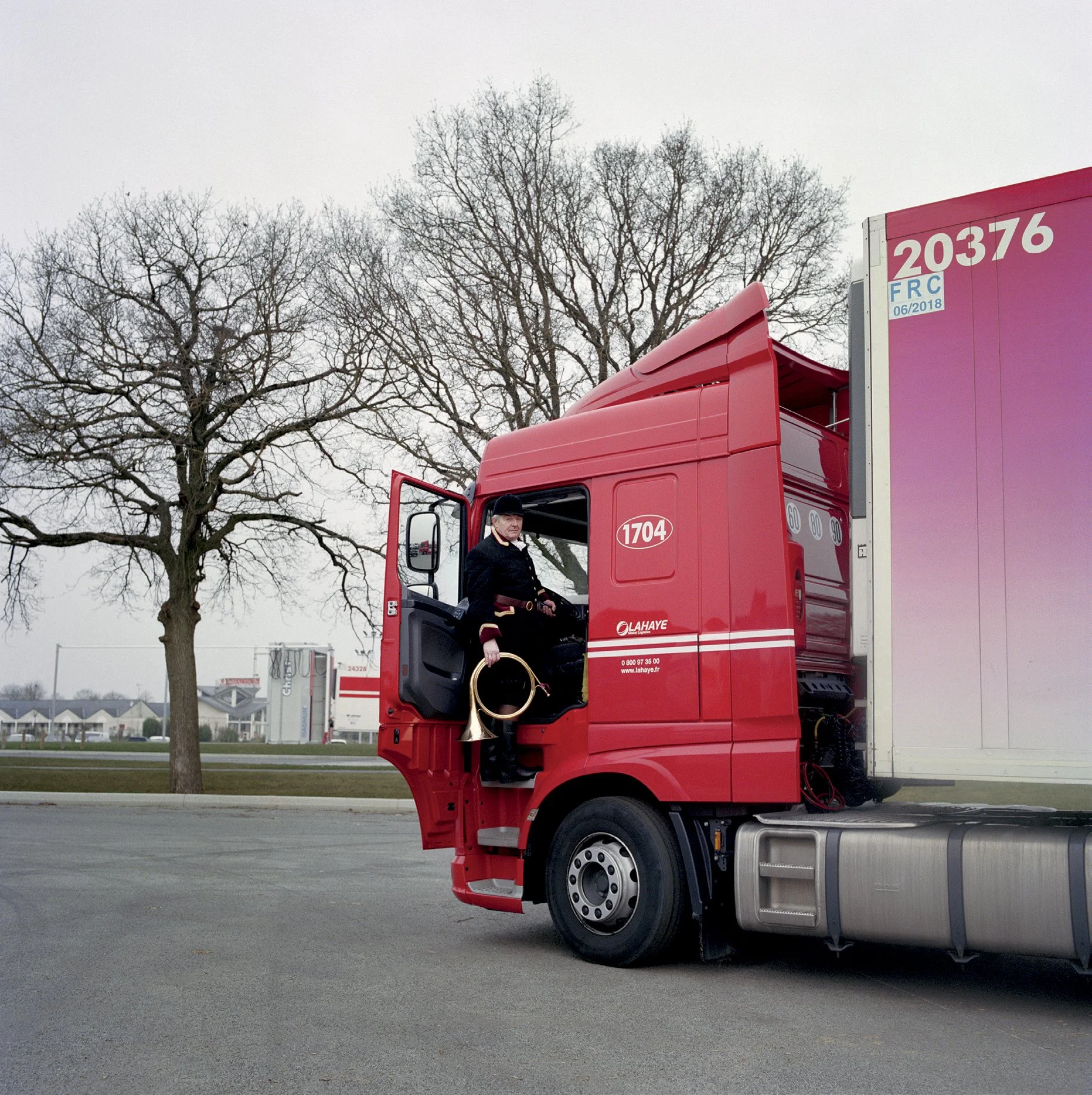 A man in traditional uniform sits in the cab of a large red truck, holding a trumpet. Bare trees in background, cloudy sky.