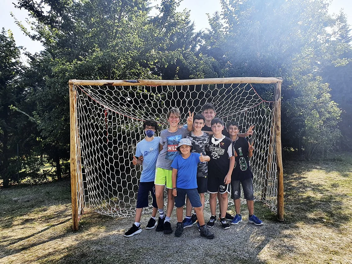 Group of eight smiling children making peace signs in front of a wooden soccer goal with net, outdoors with trees in the background.