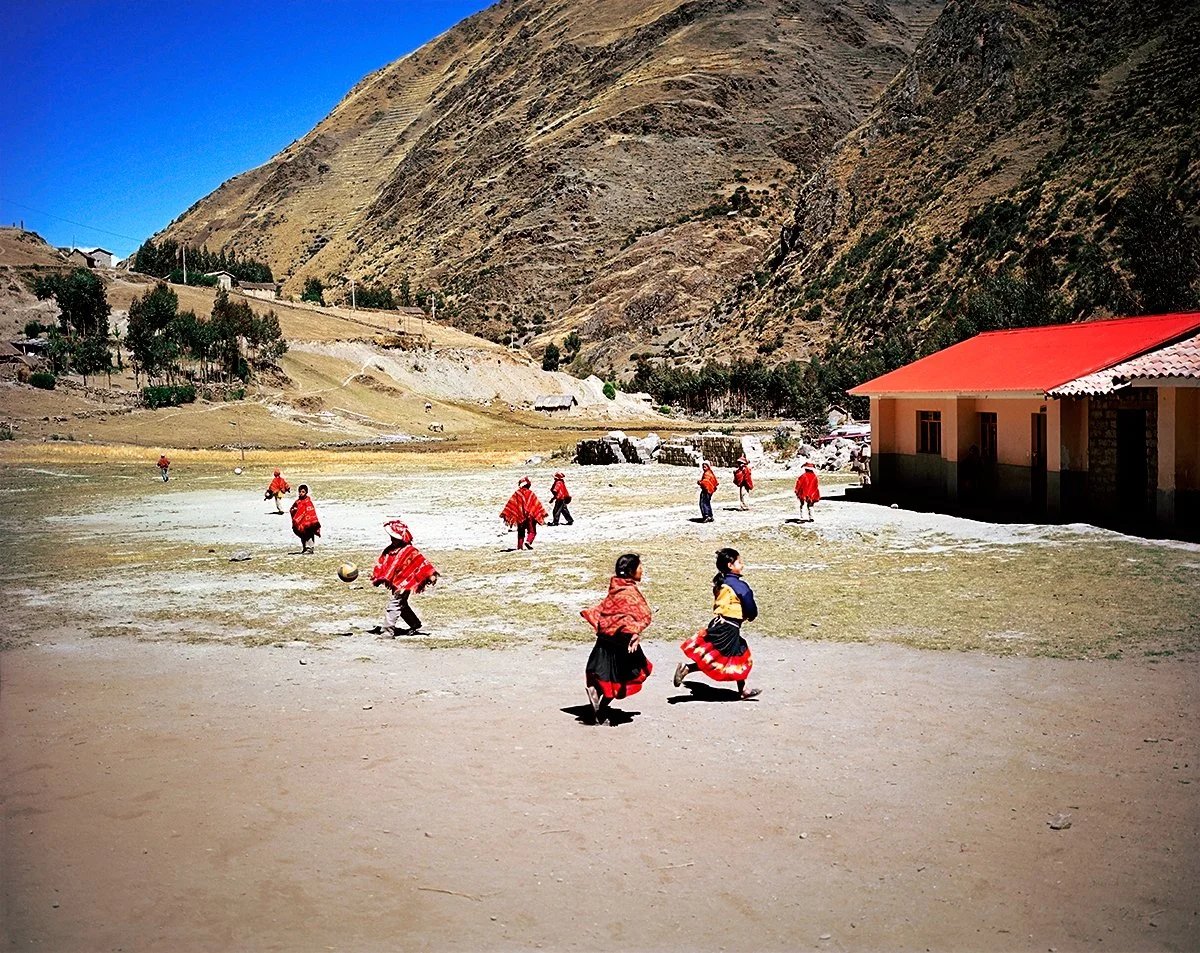 Children play ball in an open-air courtyard, dressed in colorful traditional clothing, with a mountain in the background and a red-roofed house.