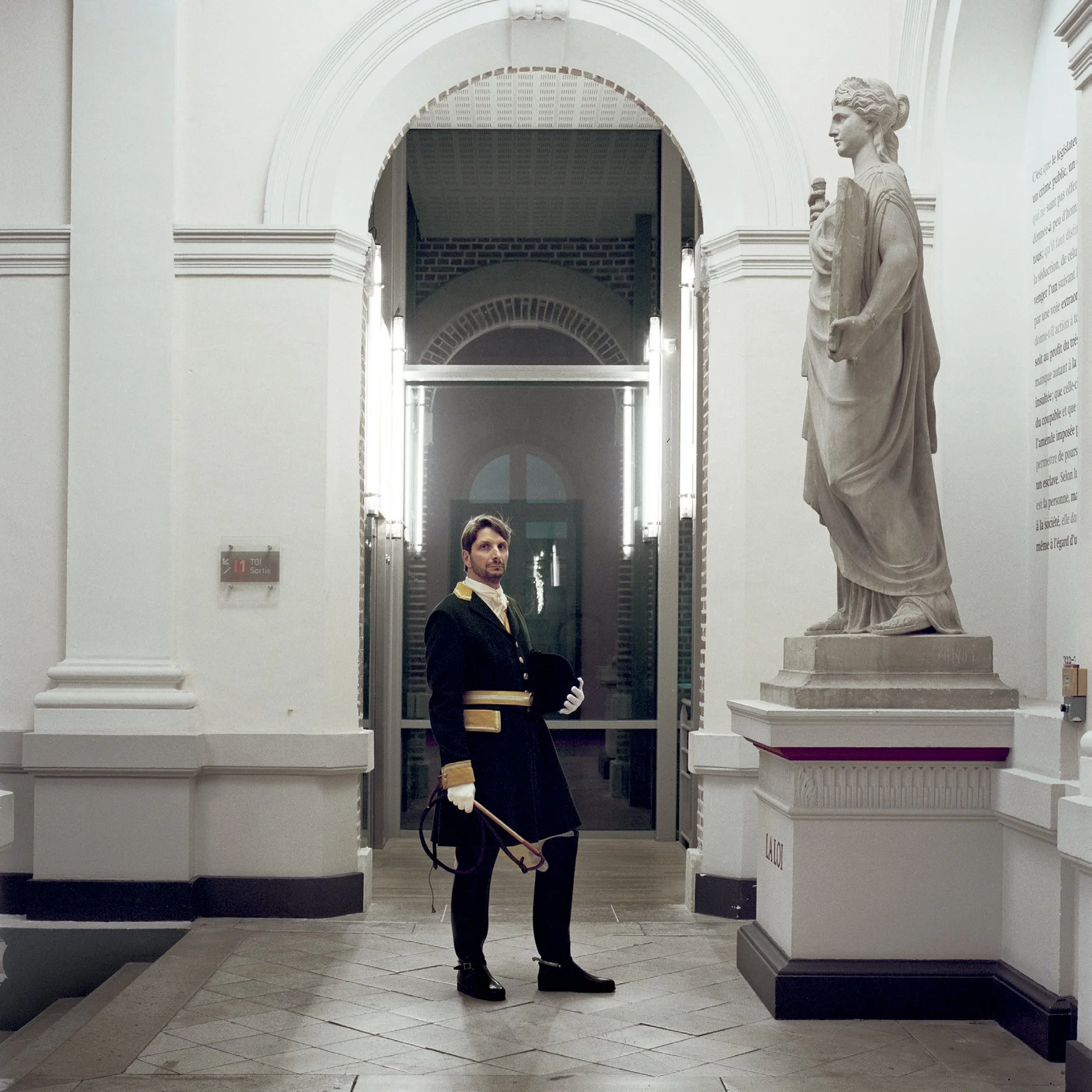 A man dressed in a historical uniform in a museum, posing next to a classic marble sculpture in a room of elegant old design.