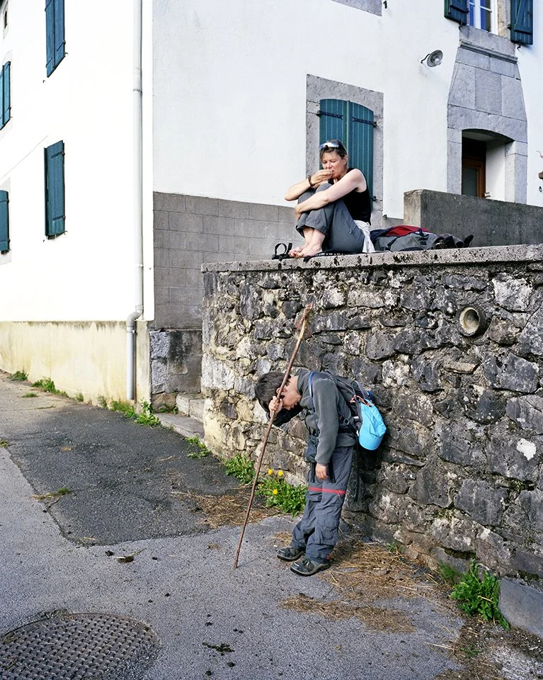 A woman sitting on a stone wall gazing pensively, a boy picking up an object or rummaging near the ground, in an urban environment with buildings and a paved street.