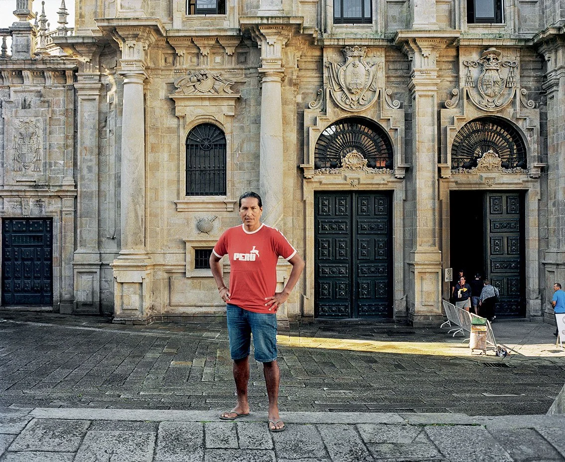 A man in a red T-shirt and denim shorts stands in front of the stone facade of a historic building with massive black doors and carved ornamentation.