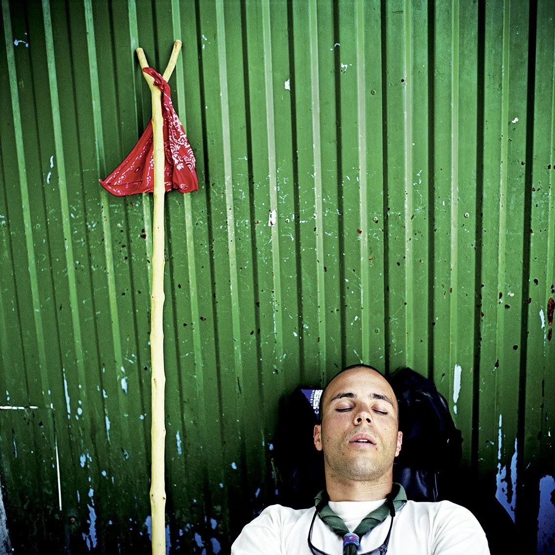 A man lies, eyes closed, against a green metal wall, with a wooden stick beside him. The stick has a red bandana on it.