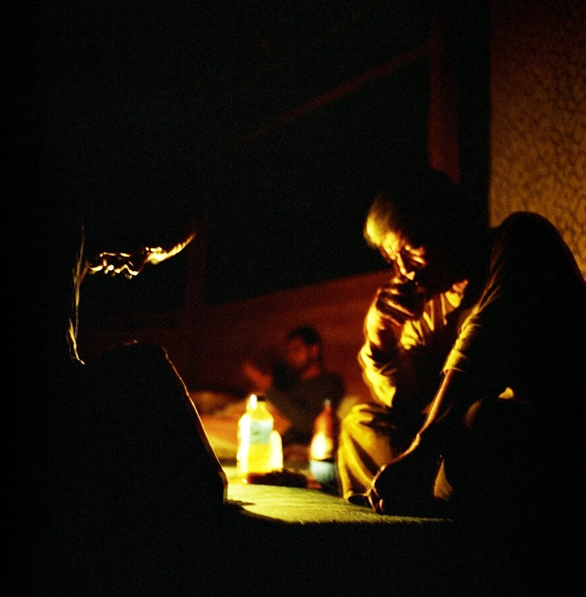 A person thinking or chatting in a dark room, lit by an orange light, with other people blurred in the background.