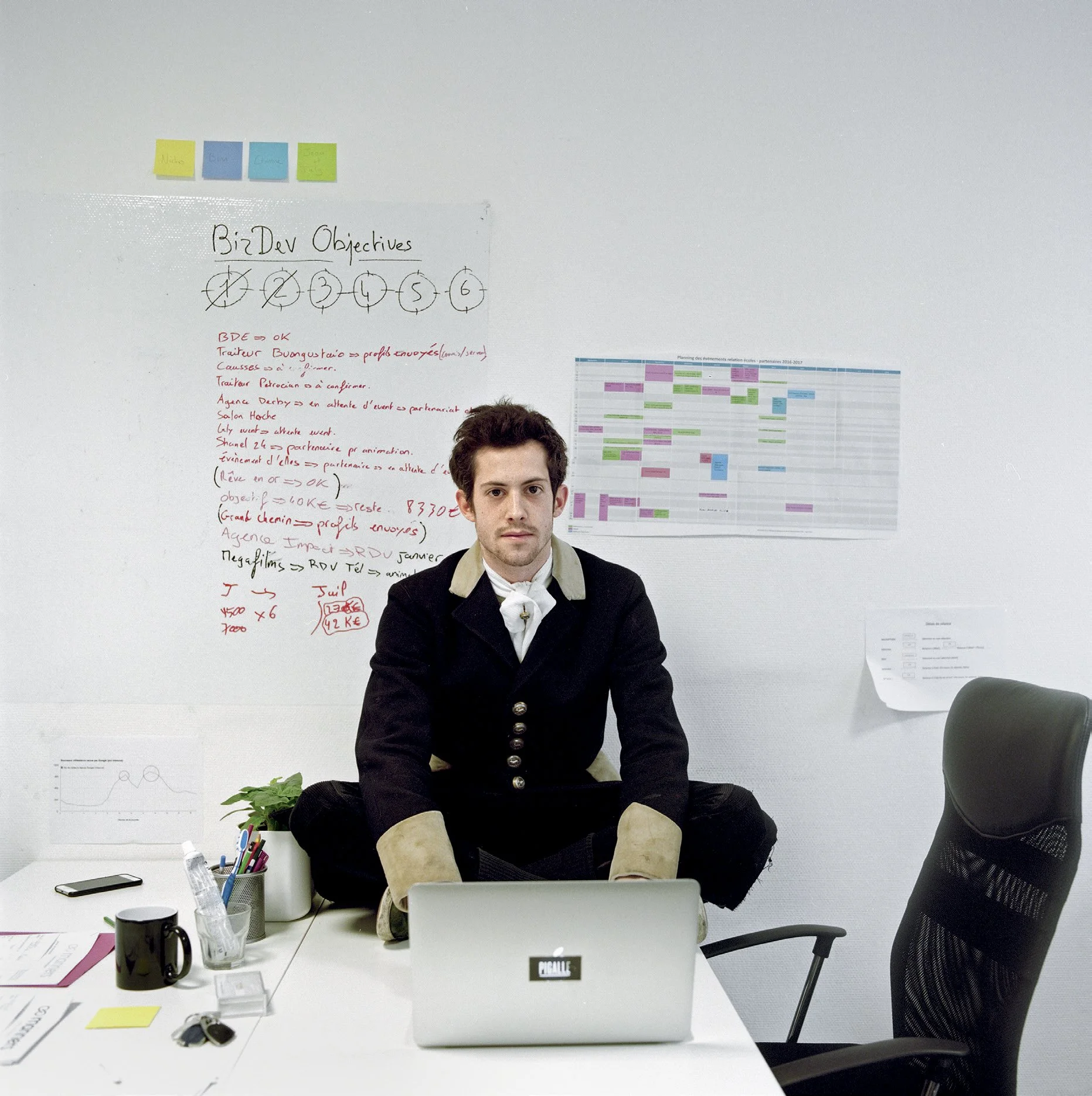 A man sitting in the center of a white desk with a laptop in front of him, notes, a mug, a plant and various objects on the table, with a whiteboard and graphics on the wall behind him.