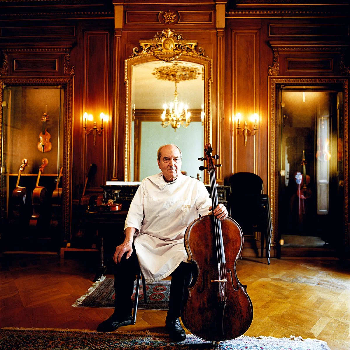 An elderly man dressed as a doctor or chef in an elegant wooden room with a large mirror, playing the cello.